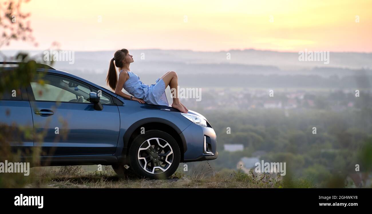 Happy young woman driver in blue dress laying on her car hood enjoying warm summer day. Travelling and vacation concept. Stock Photo