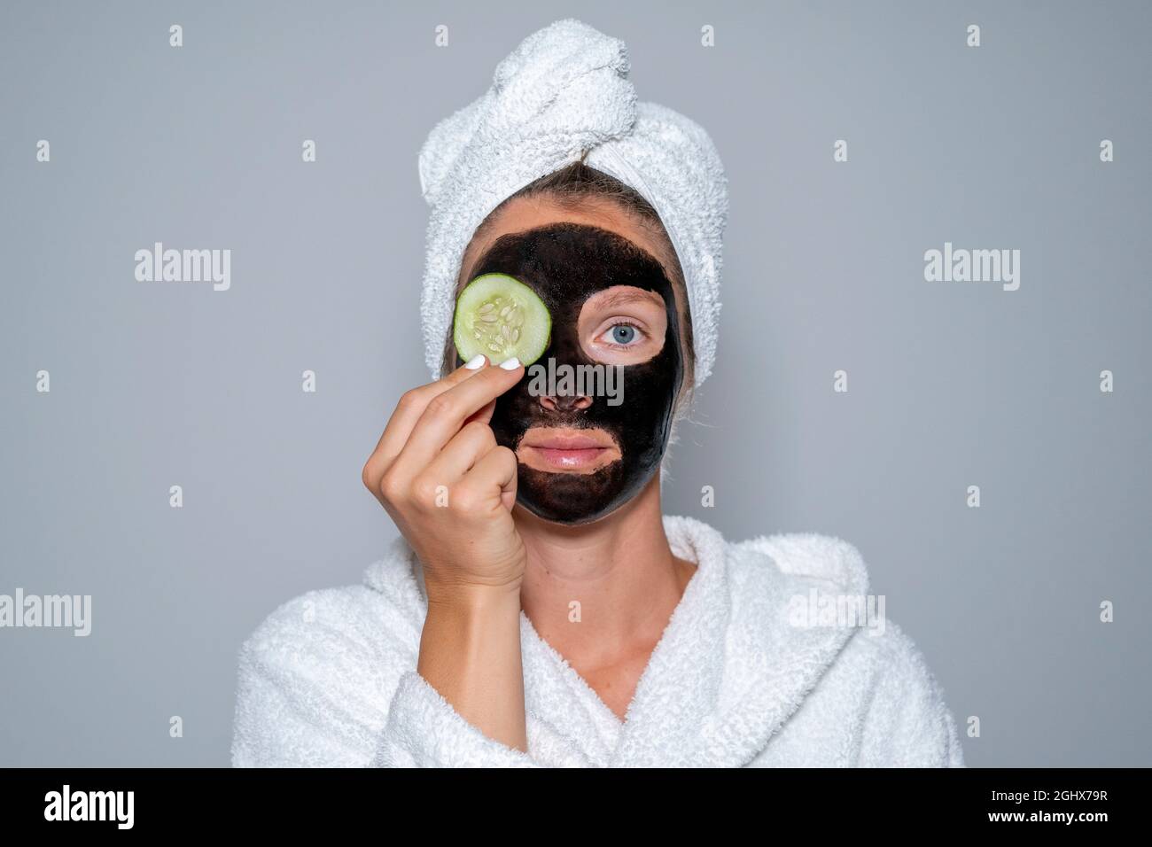 Young woman at home spa with black facial mask grabbing a cucumber slice. Wellness concept face treatment Stock Photo