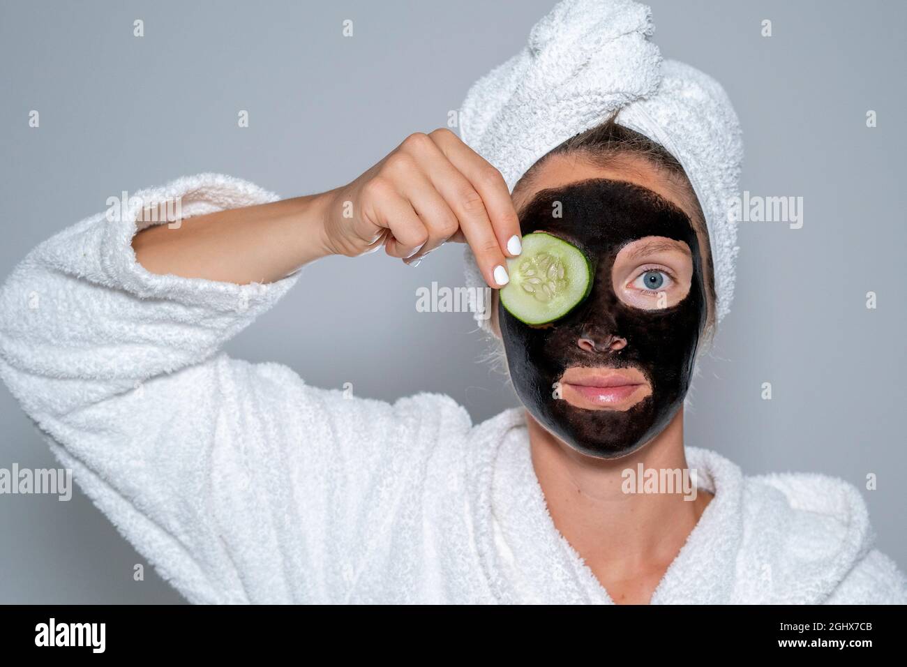 Young woman at home spa with black facial mask grabbing a cucumber slice. Wellness concept face treatment Stock Photo