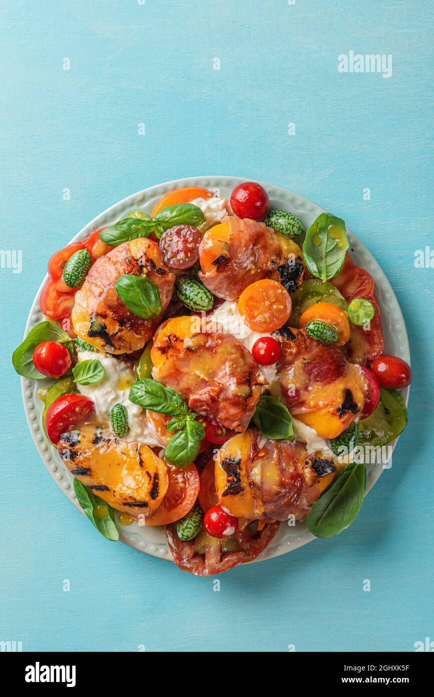 Delicious salad with burrata cheese, grilled peaches and prosciutto, burrata cheese, tomatoes, cucumber, olive oil and basil. Top view. Healthy diet f Stock Photo
