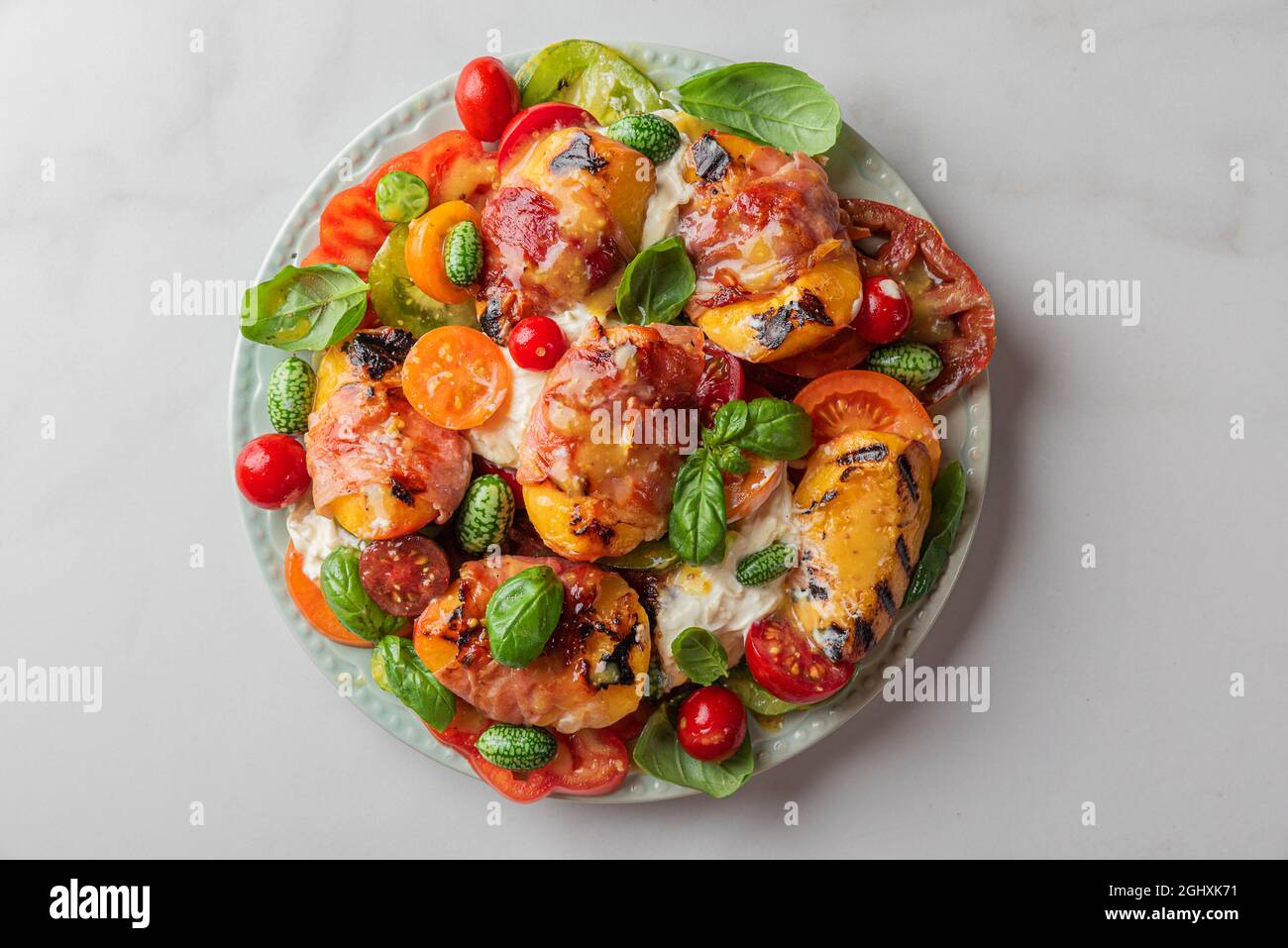 Grilled peaches and prosciutto salad with burrata cheese, tomatoes, cucumber and basil in a plate on white table. Top view. Healthy diet food concept Stock Photo