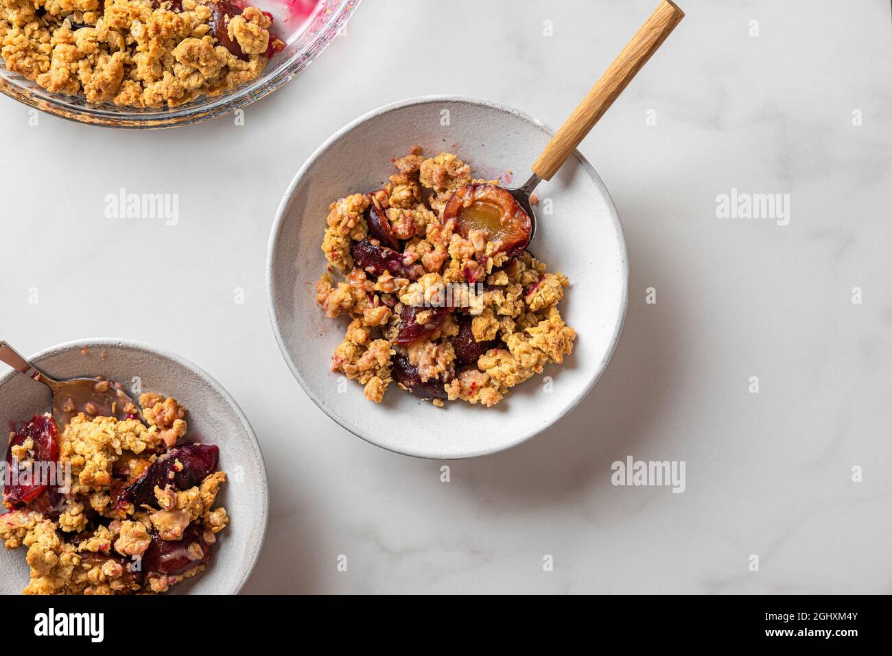 Fruit crumble. Plum crumble pie in plates with spoons on white marble