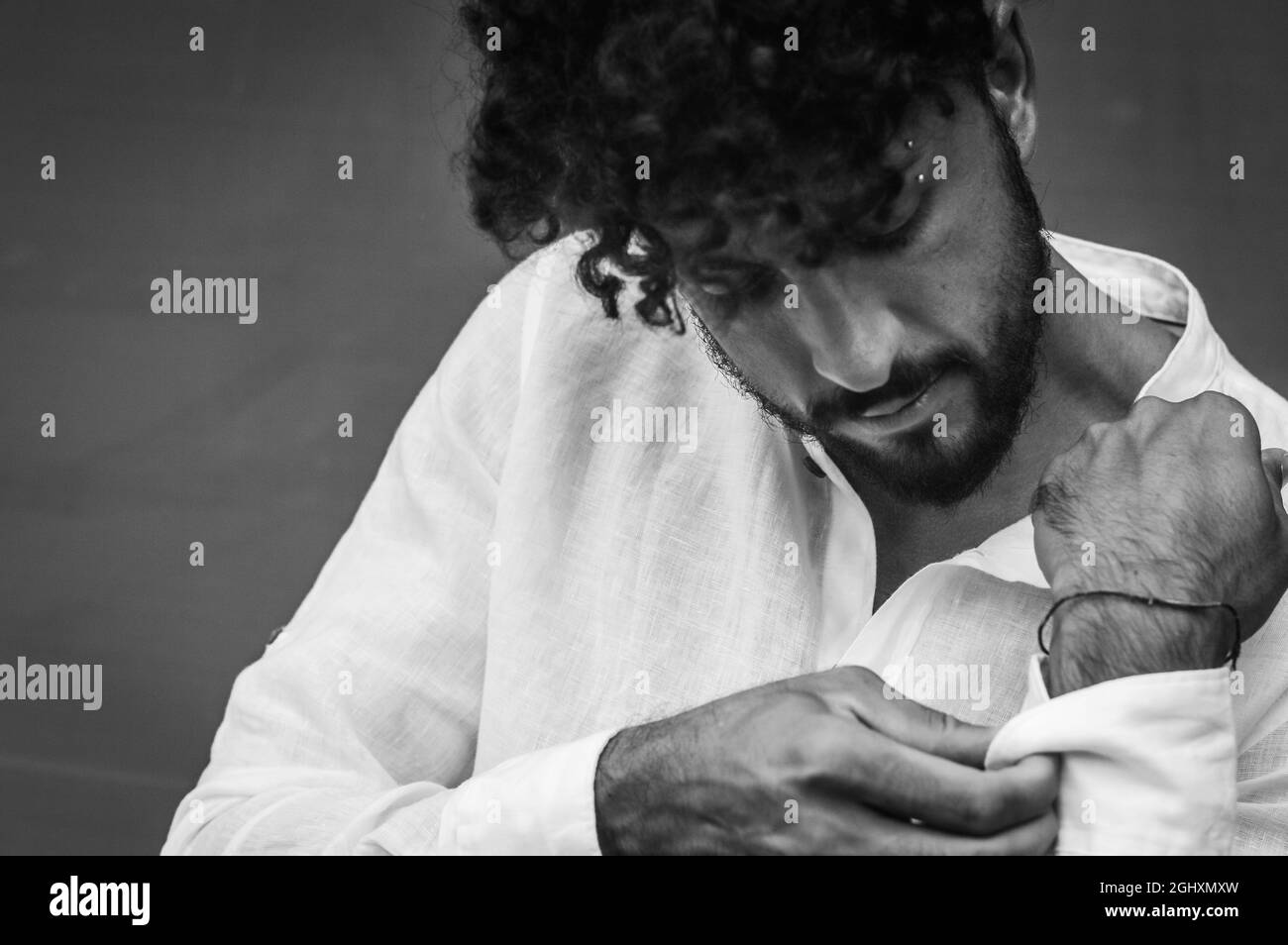 black and white portrait of a young man with a beard and curly hair adjusting the sleeves of his white shirt while dressing Stock Photo