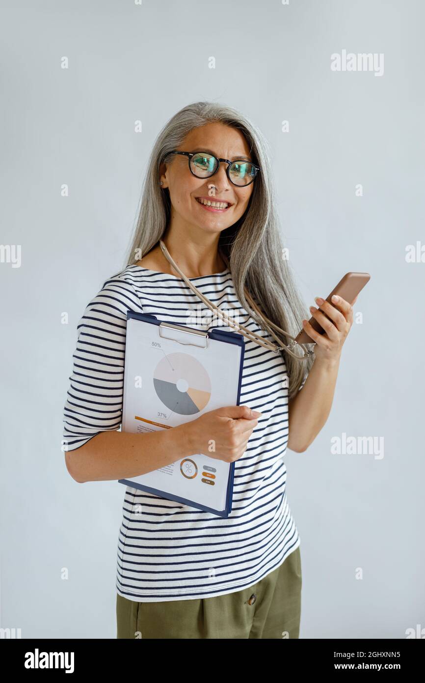 Silver haired Asian lady with glasses holds colorful diagrams and smart phone in studio Stock Photo