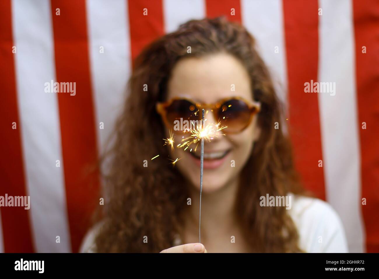 Blurred Young redhead Hispanic and Caucasian woman holding a sparkler in focus standing in front of the American flag. Stock Photo