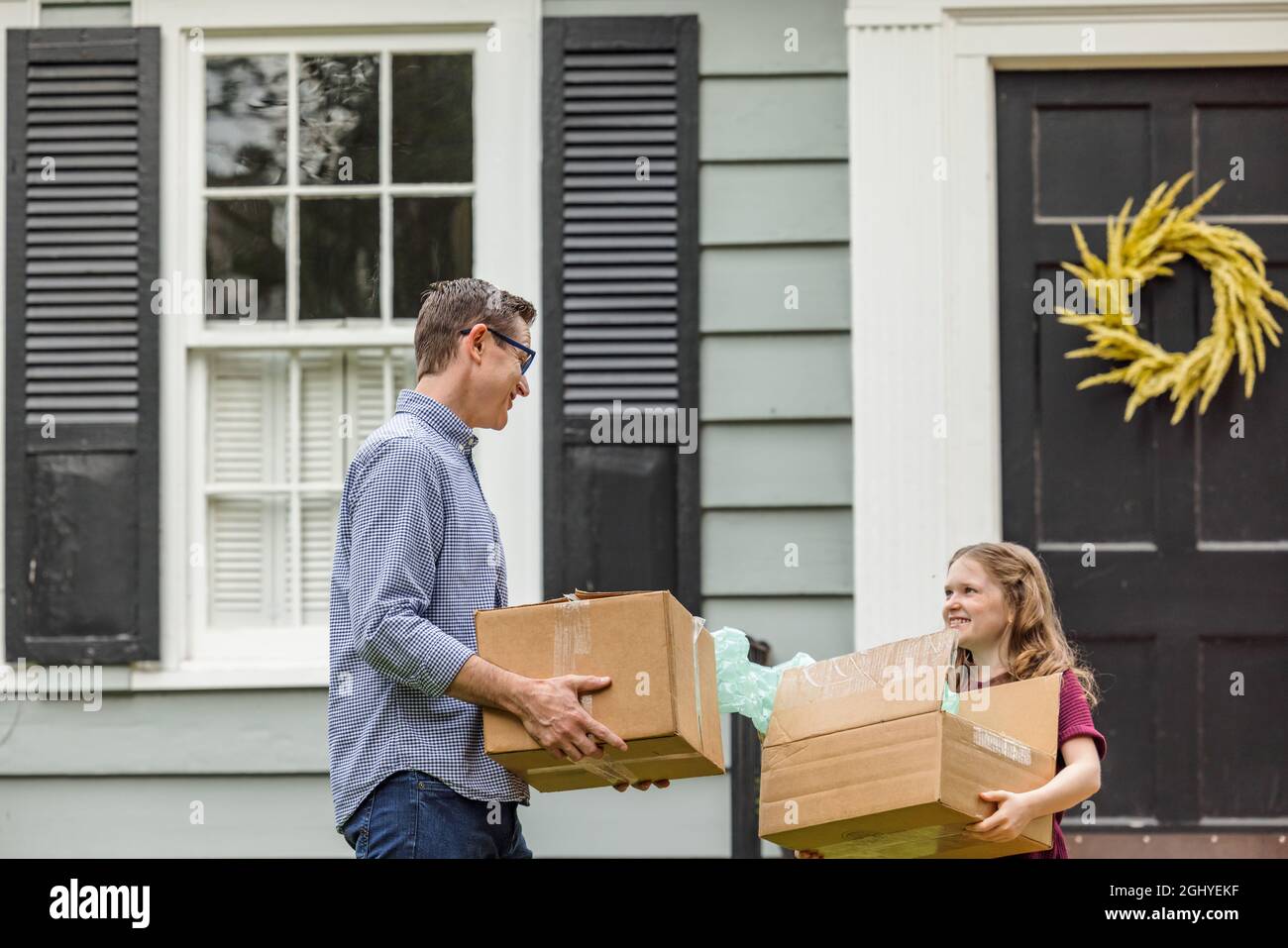 A father and daughter holding moving boxes outside a small blue cottage house getting ready for a move Stock Photo