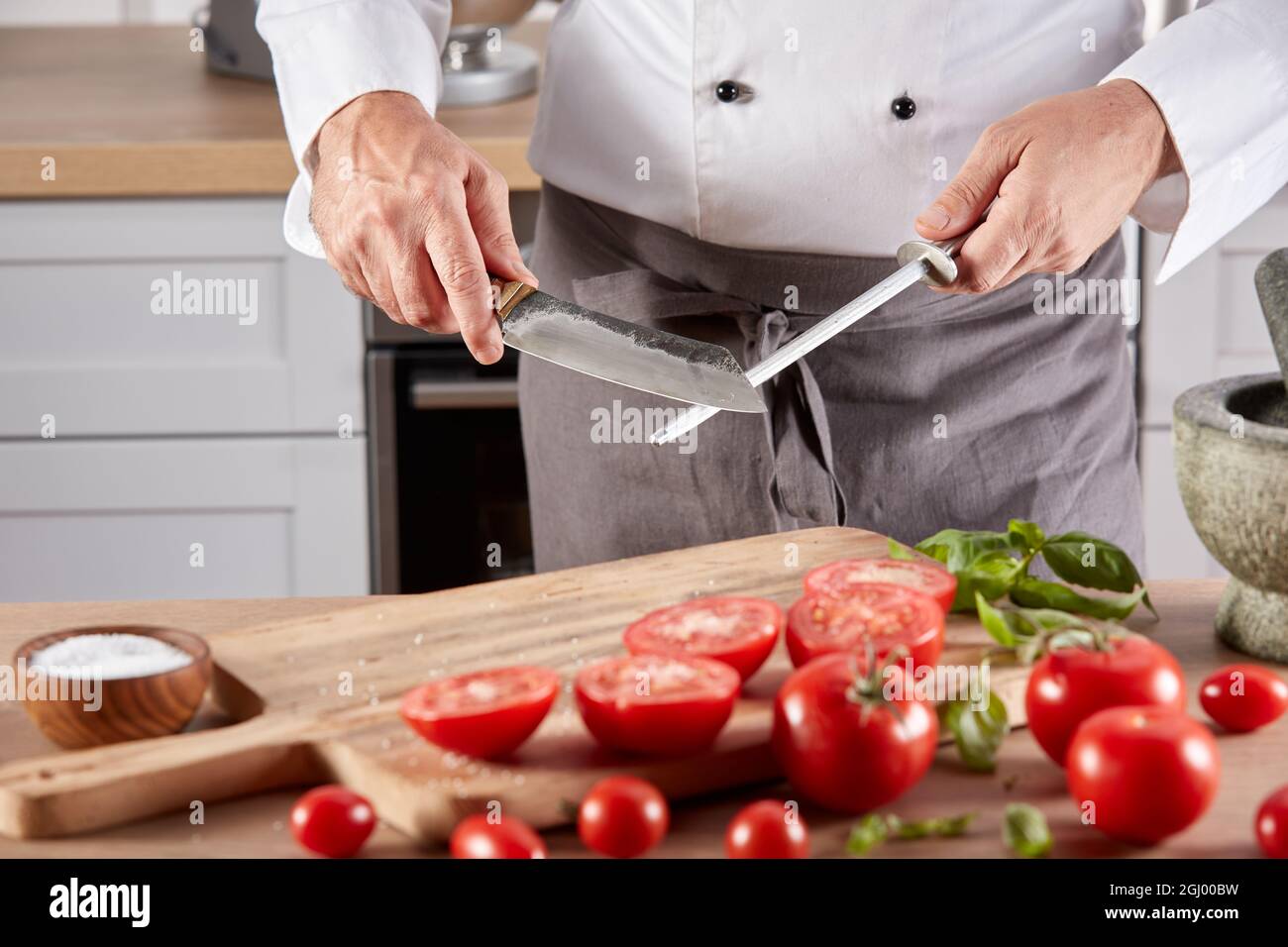 Anonymous crop male chef sharpening blade of knife while standing at wooden table with tomatoes before cooking in kitchen Stock Photo