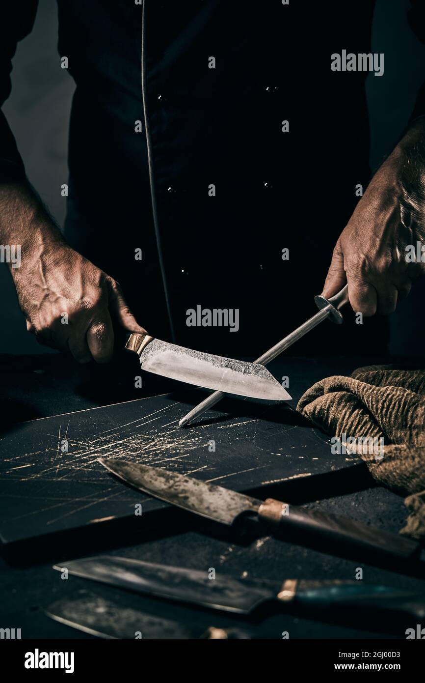 Crop faceless male chef sharpening metal blade of knife with sharpener while working in dark kitchen Stock Photo