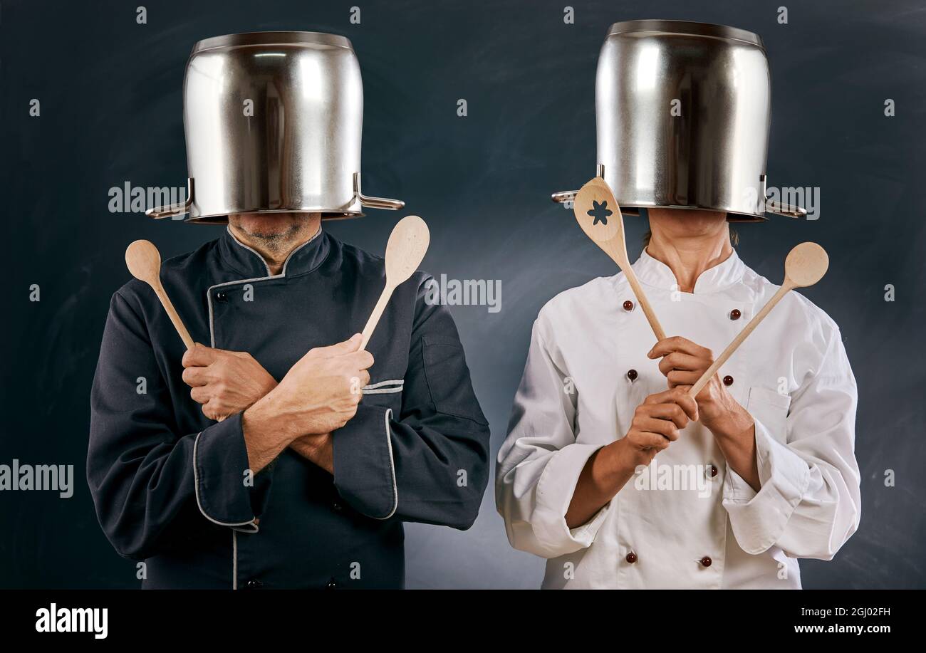 Two male chefs in uniform spoofing around with large stainless steel pots upended over their heads and holding crossed wooden spoons over a dark backg Stock Photo
