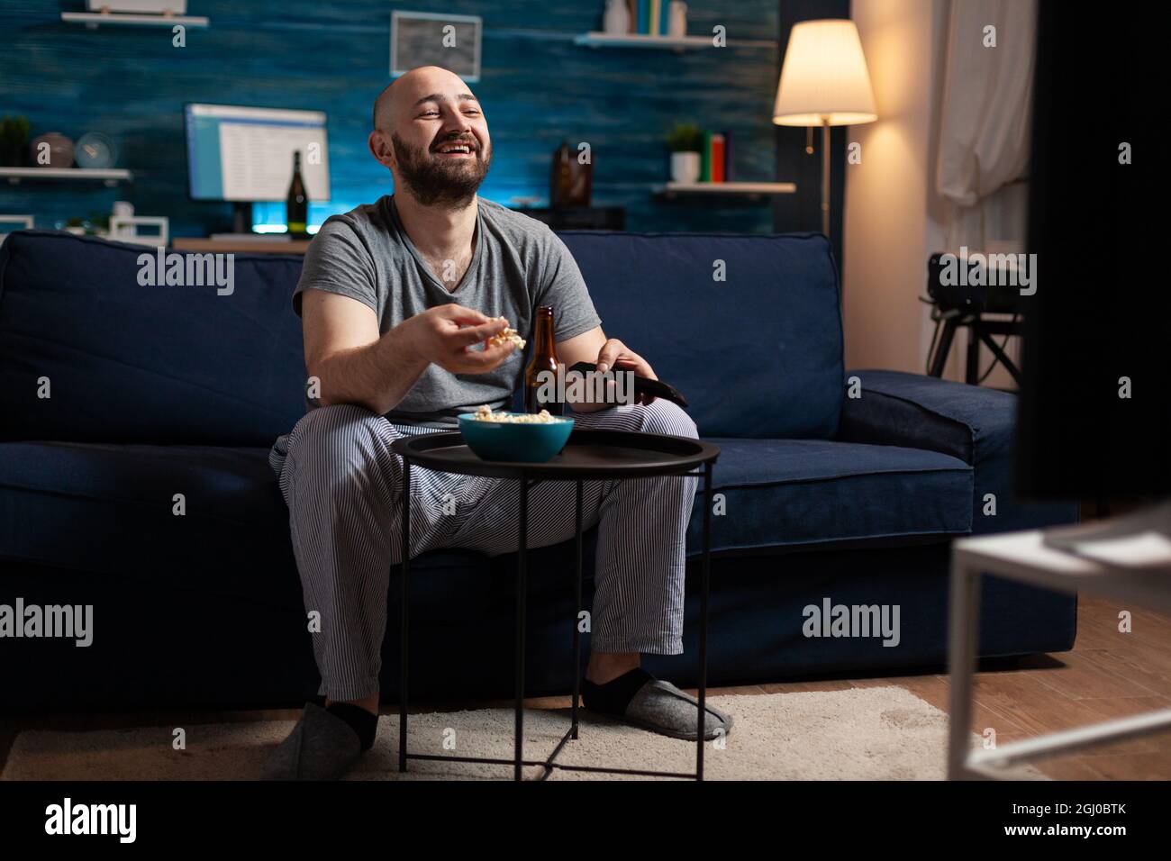 Man enjoying relax time watching tv comedy series at home sitting on comfortable couch dressed in pajamas eating popcorn. Excited amused home alone male relaxing on cozy sofa in living room late night Stock Photo