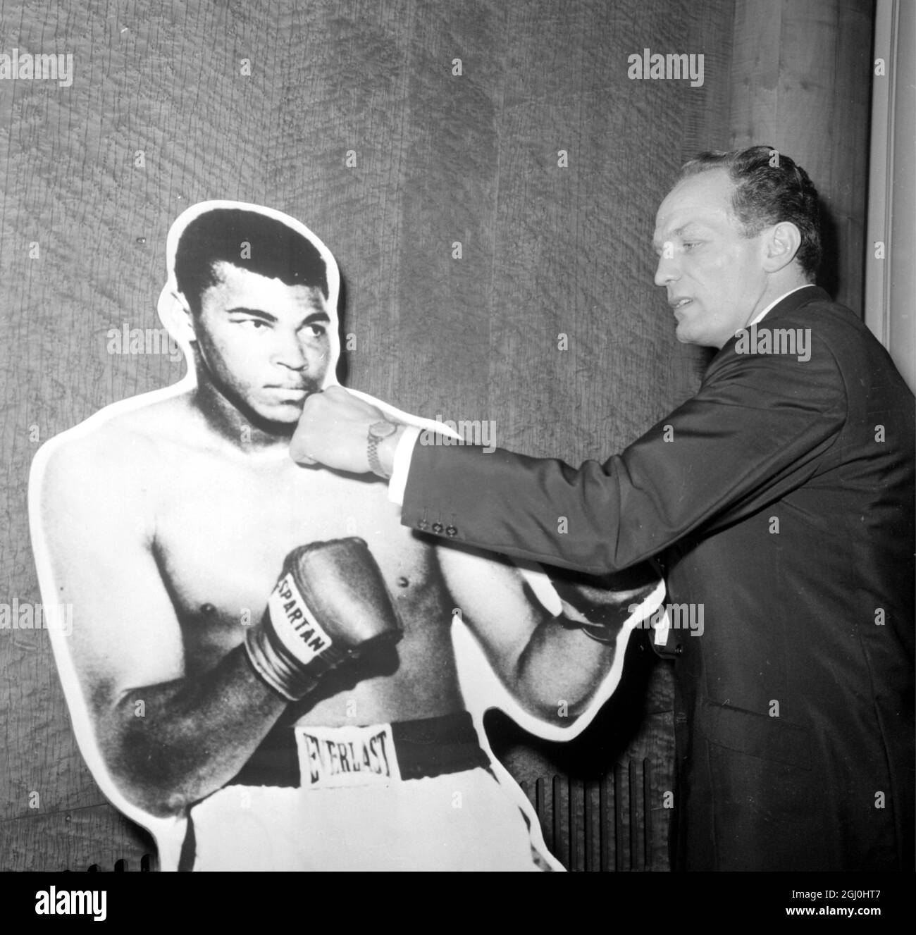 London: Aiming a playful punch at this blown-up picture of World Heavyweight champion Cassius Clay today is British Heavyweight Champion Henry Cooper, pictured at the Odeon, Leicester Square, shortly after signing a contract to fight Clay in London on May 21. The title bout will take place at the Arsenal Football Club Stadium, Highbury, London. 19 April 1966 Stock Photo