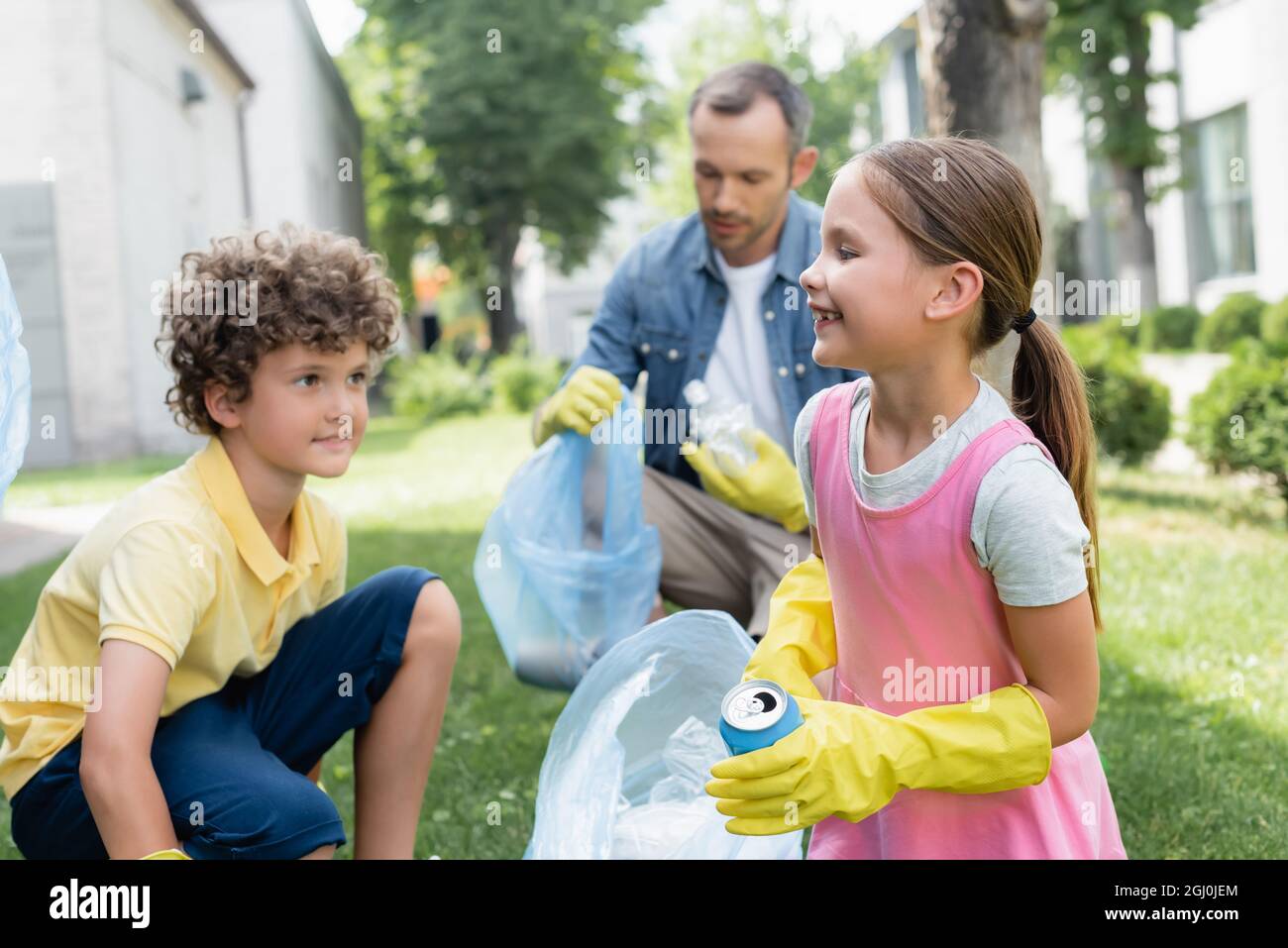 Happy kid holding tin can and trash bag near blurred brother and dad