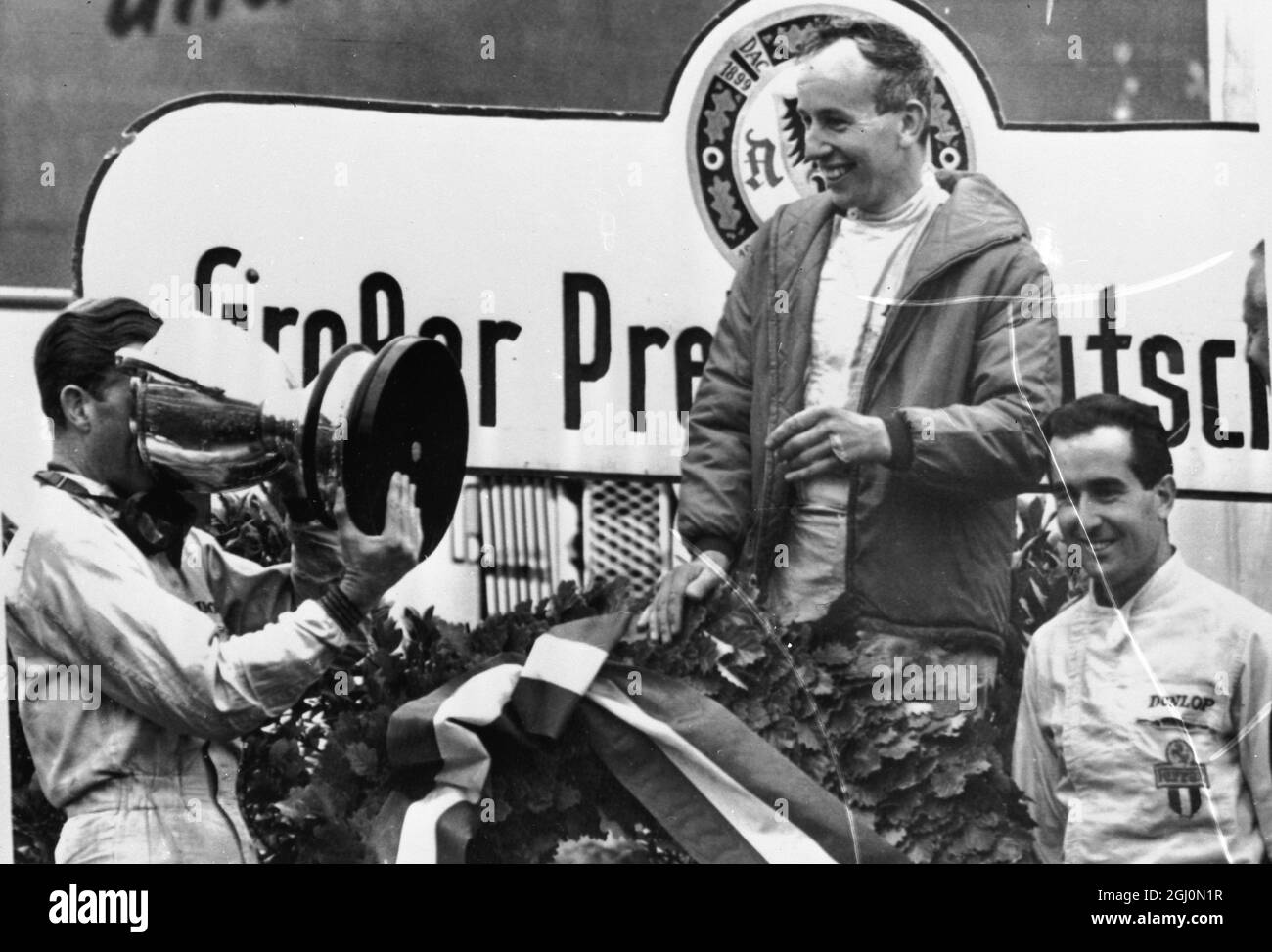 John Surtees looks smilingly on as Graham Hill drinks Champagne out of the Gian Cup presented to Surtees after he had won the German Grand Prix at Nurburgring. On the right is Lorenzo Bandini. 1964 Stock Photo