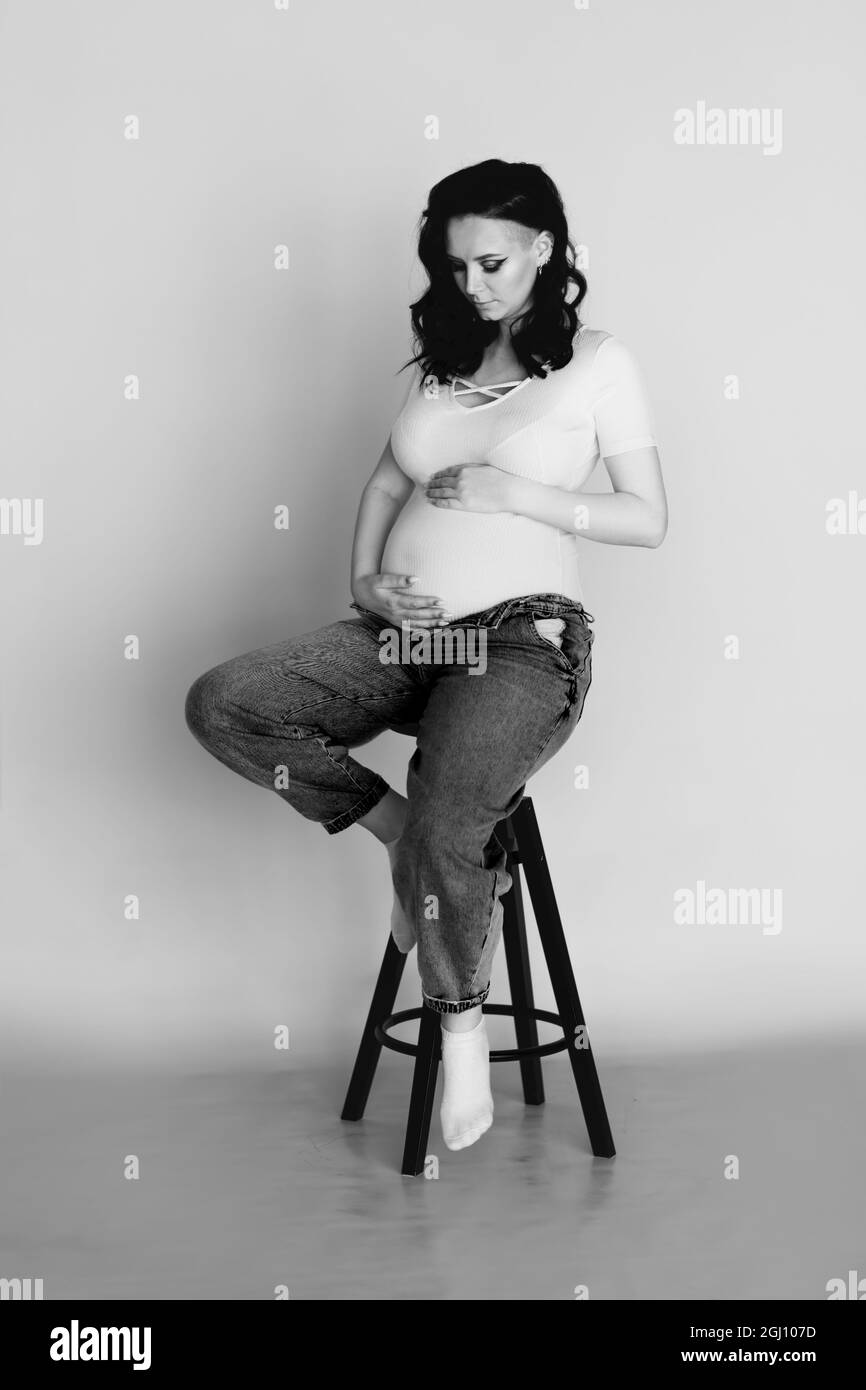 Black and white portrait of pregnant woman wearing jeans and white T-shirt and sitting on chair on beige background. Pregnancy, motherhood and happine Stock Photo