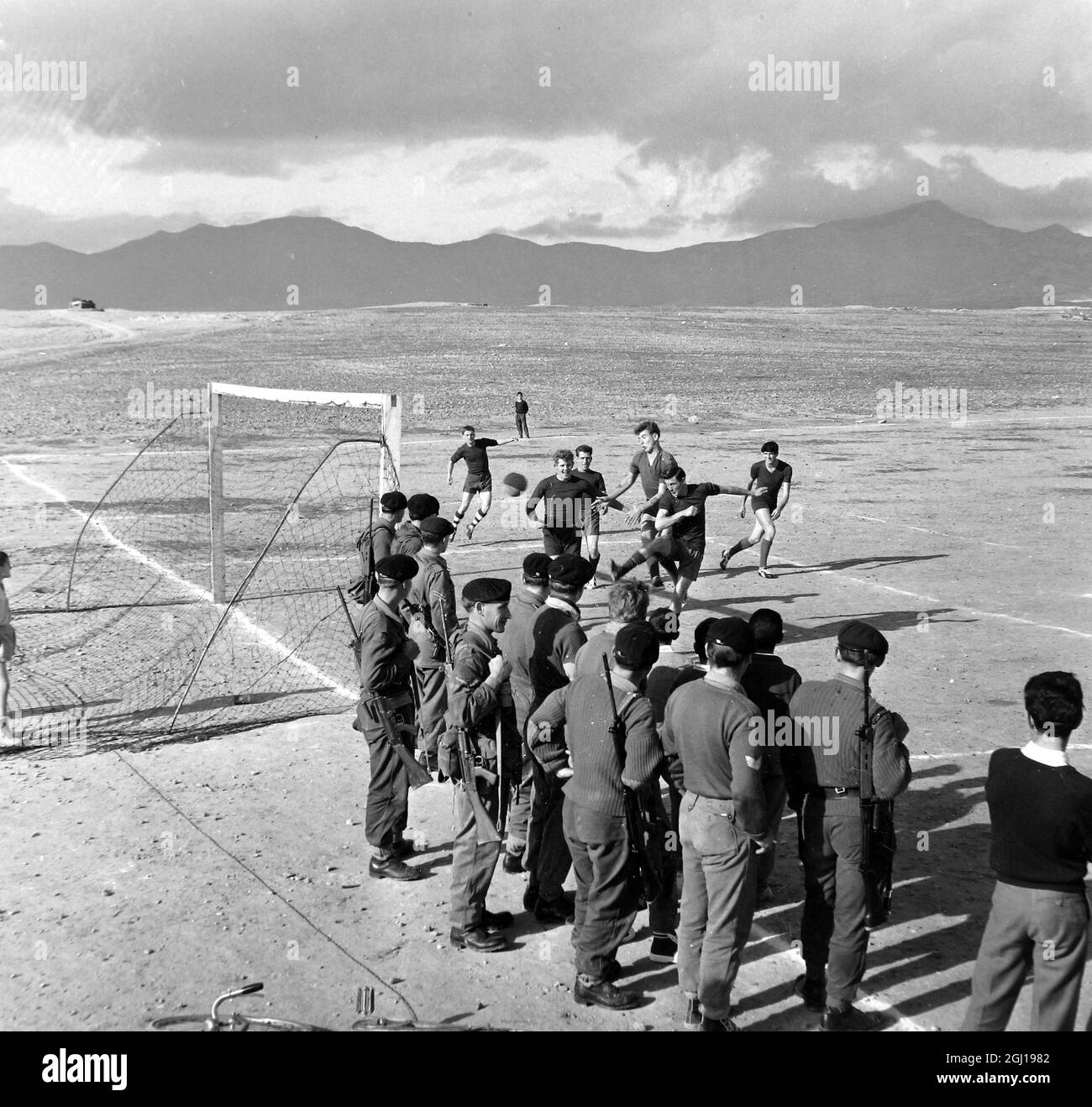 16 FEBRUARY 1964 BRITISH ARMY TROOPS OF THE GLOUCESTERSHIRE REGIMENT, WITH GUNS SLUNG ACROSS THEIR SHOULDERS, WATCH FELLOW COMRADES PLAY FOOTBALL IN NICOSIA, CYPRUS Stock Photo