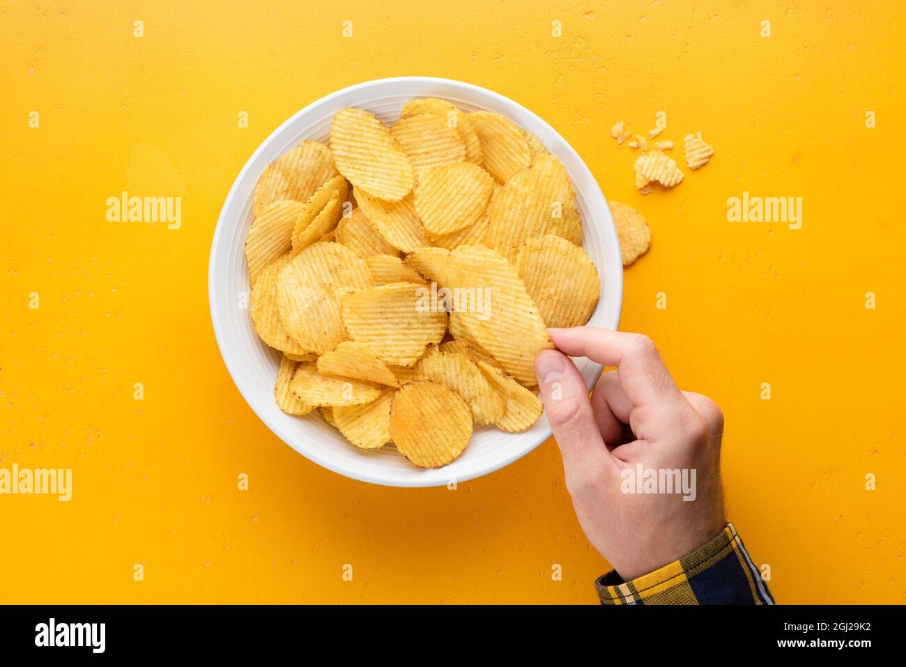 Spicy potato crisps in a bowl on yellow background. Male hand picking potato crisps. Junk food, unhealthy eating, party food concept Stock Photo