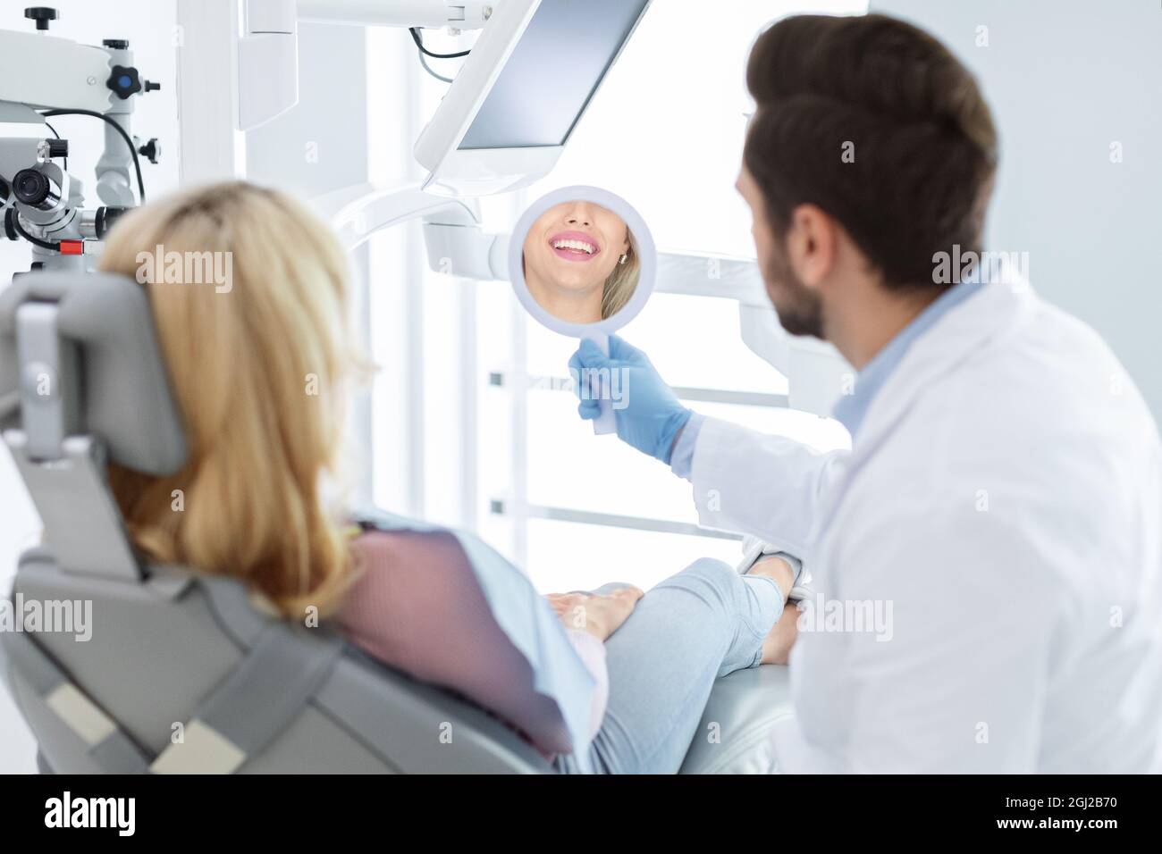 Bearded man dentist holding mirror for smiling blonde woman patient, stomatologist and young lady checking results of treatment, healthy white smile r Stock Photo