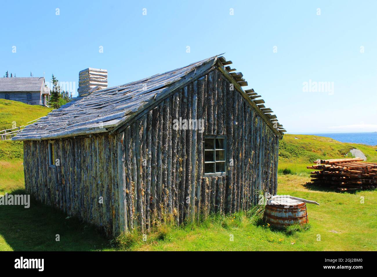 An old log house, with an old wooden church on a hill in the background, Random Passage Site, New Bonaventure, Newfoundland. Stock Photo