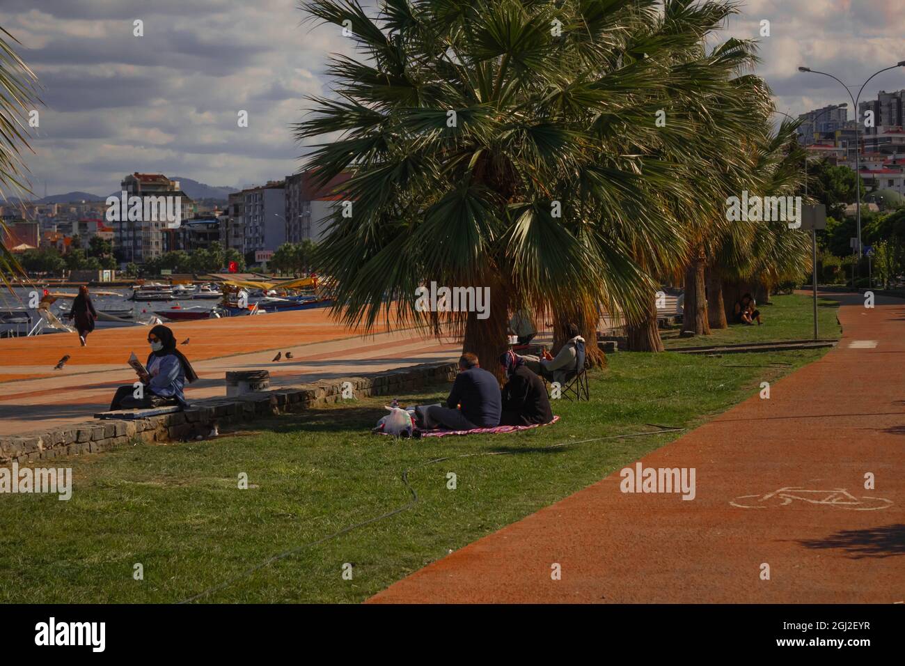 People having a picnic under palm trees. Covered woman reading a book. Walking path of the city. Stock Photo