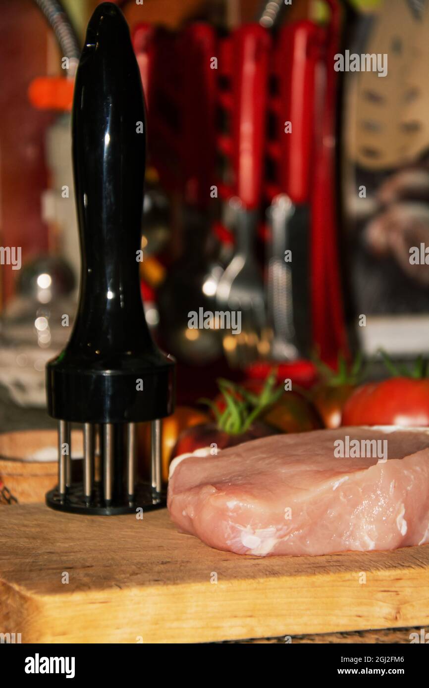 A piece of raw pork meat lies next to a meat softener on a wooden cutting board on the kitchen table. In the background are red tomatoes, salt in a wooden bowl and kitchen utensils. High quality photo Stock Photo