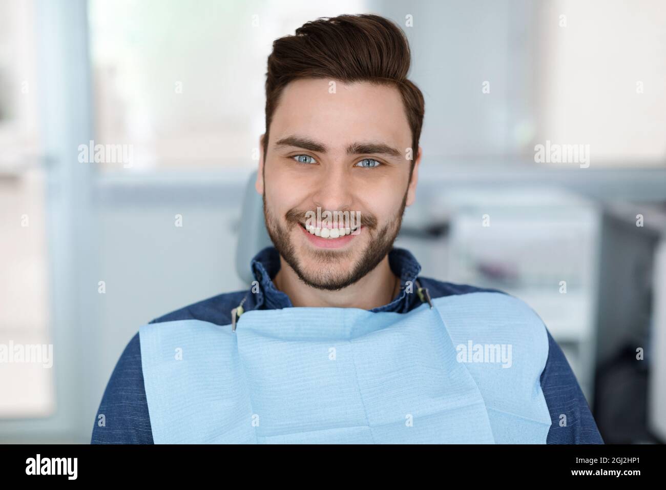 Cheerful bearde man patient sitting in dental chair and smiling at camera, copy space. Positive millennial guy showing his beautiful white smile after Stock Photo