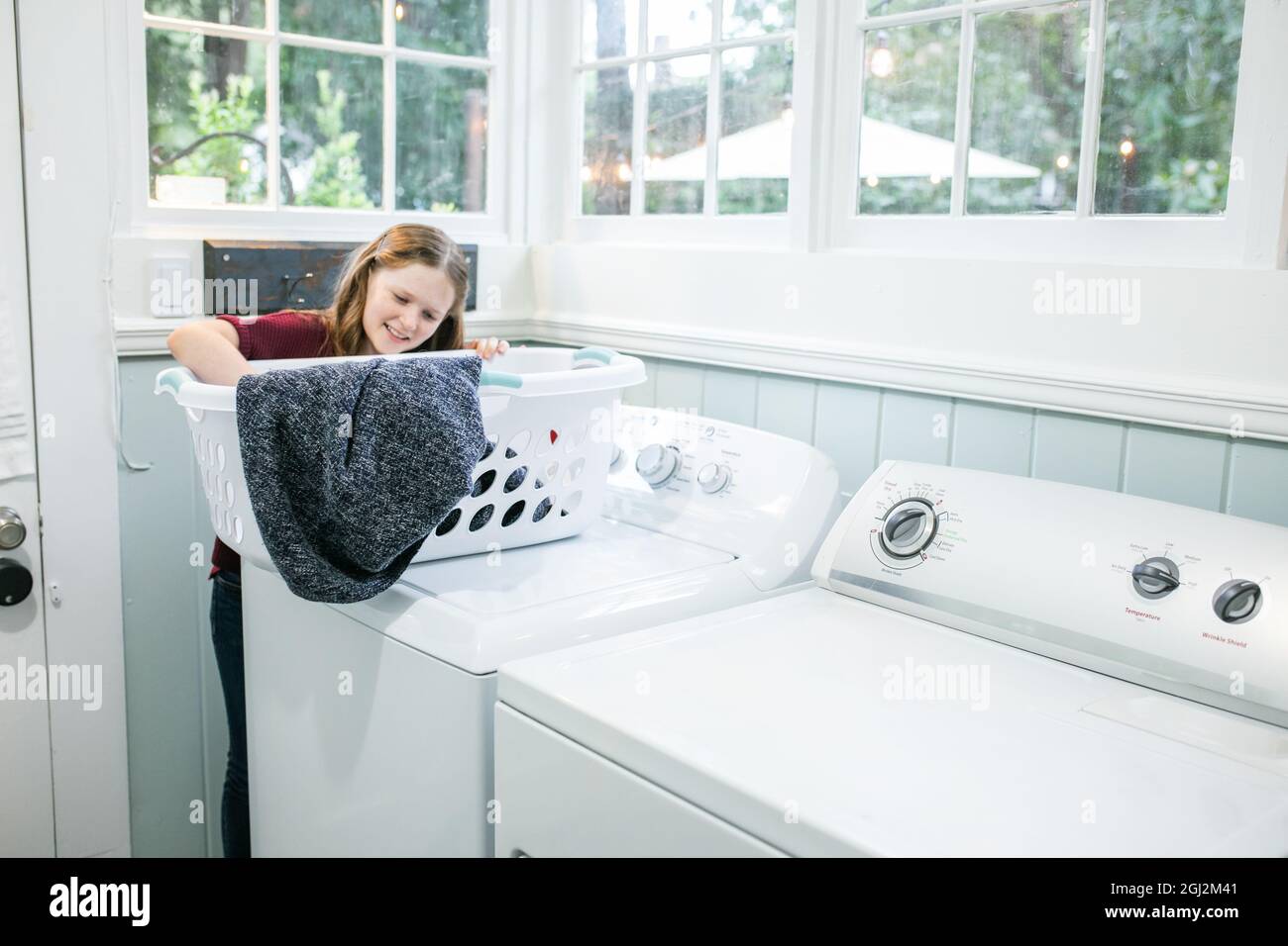 A little girl with long hair and freckles holding a laundry basket and doing the wash as laundry chores in a natural light laundry room with a washer Stock Photo