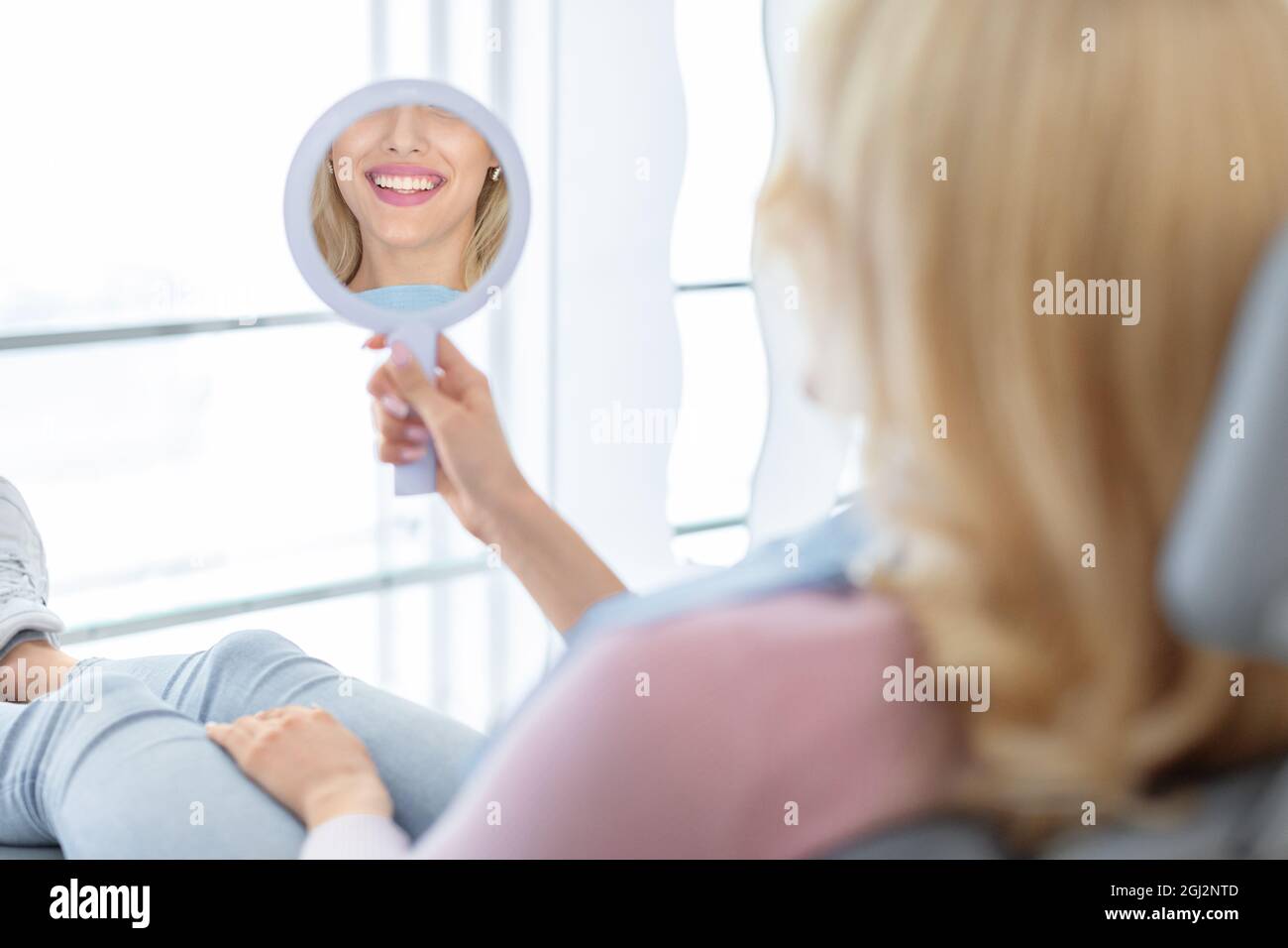 Blonde woman patient beautiful healthy white smile in dental mirror reflection, unrecognizable happy young lady checking results of treatment at moder Stock Photo