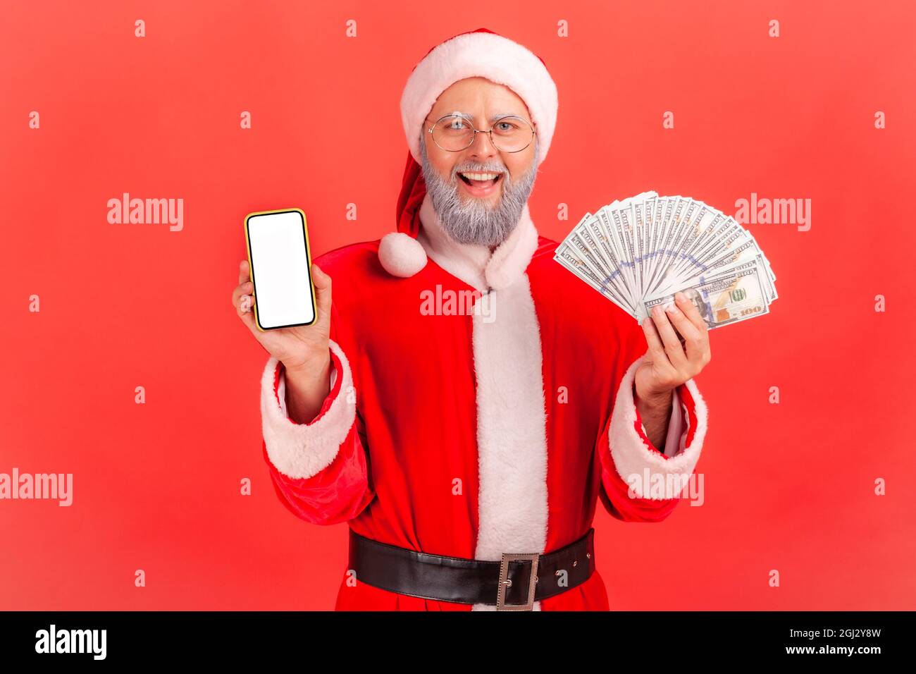 Happy smiling elderly man with gray beard wearing santa claus costume holding fan of money and showing smartphone with white blank screen. Indoor stud Stock Photo