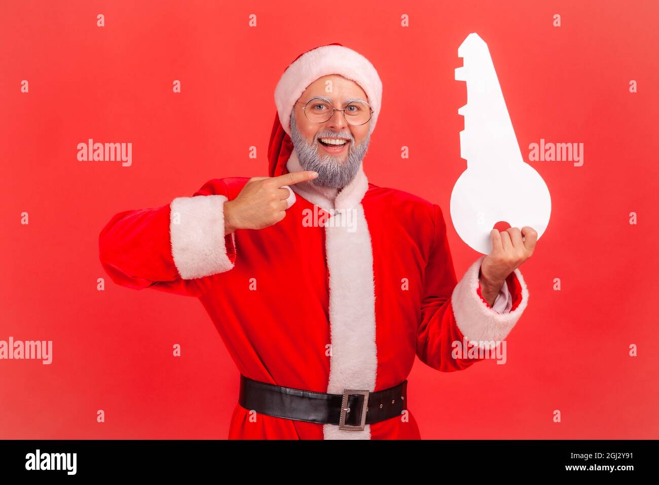 Portrait of smiling elderly man with gray beard wearing santa claus costume standing pointing to paper key, recommend agency for buying apartment. Ind Stock Photo