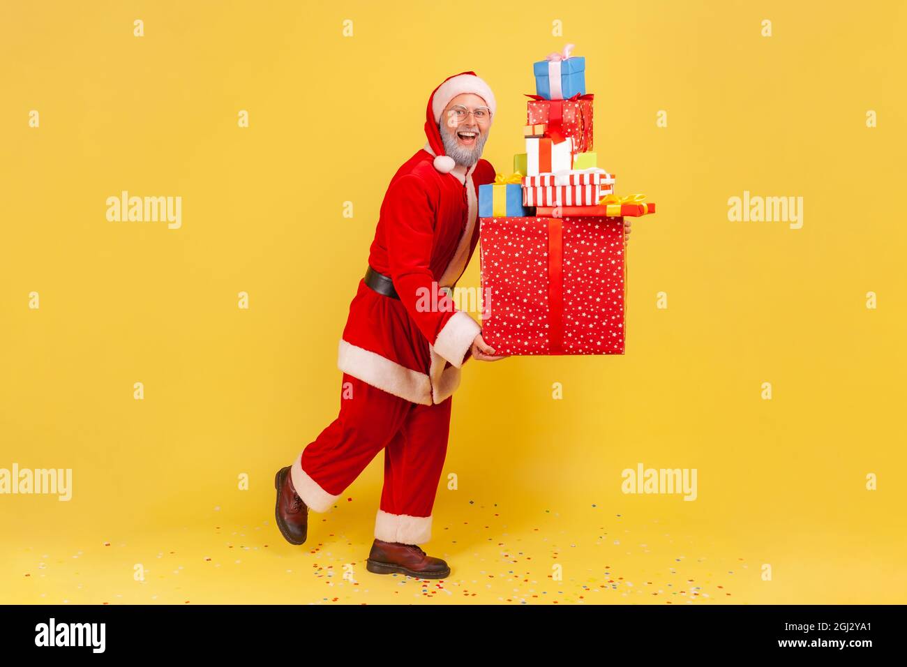 Full length portrait of elderly man with gray beard in santa claus costume holding stack of Christmas present boxes, celebrating Christmas holiday. In Stock Photo