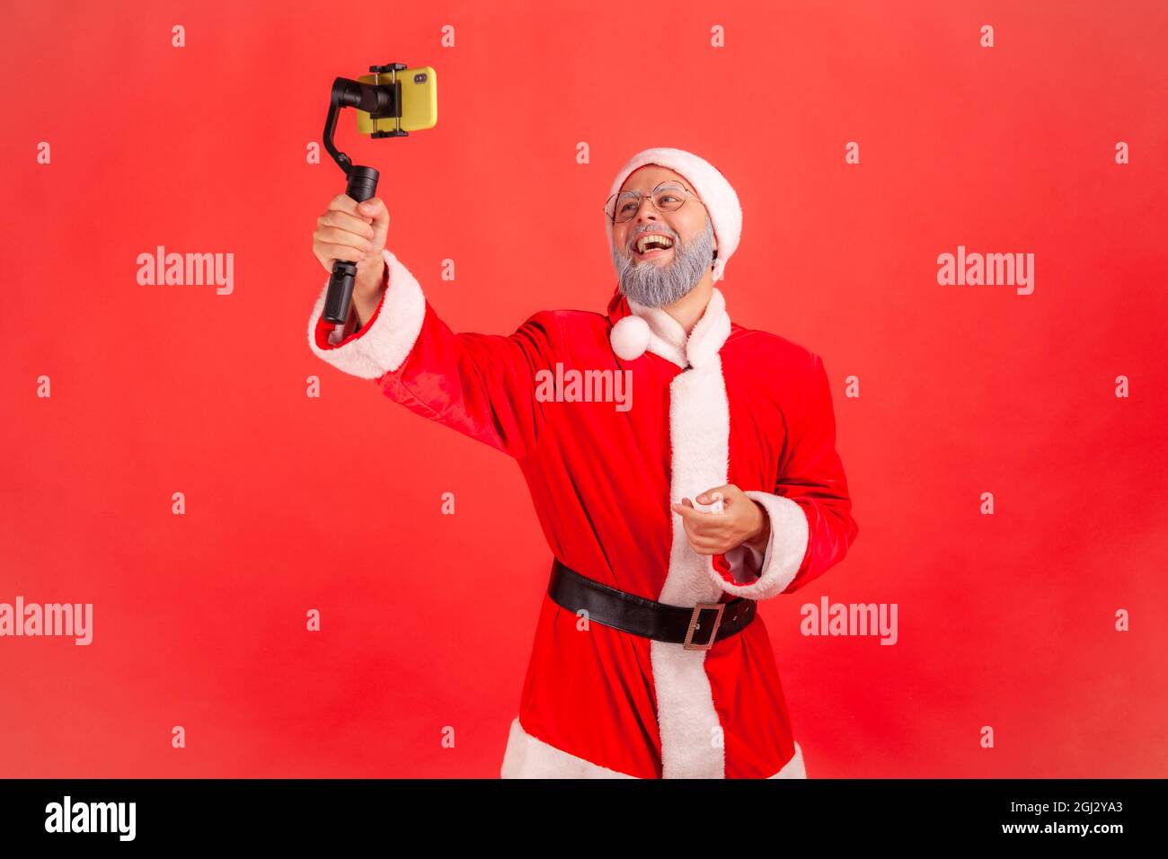 Portrait of happy smiling elderly man with gray beard wearing santa claus costume talking with followers, holding steadicam in hands, livestream. Indo Stock Photo