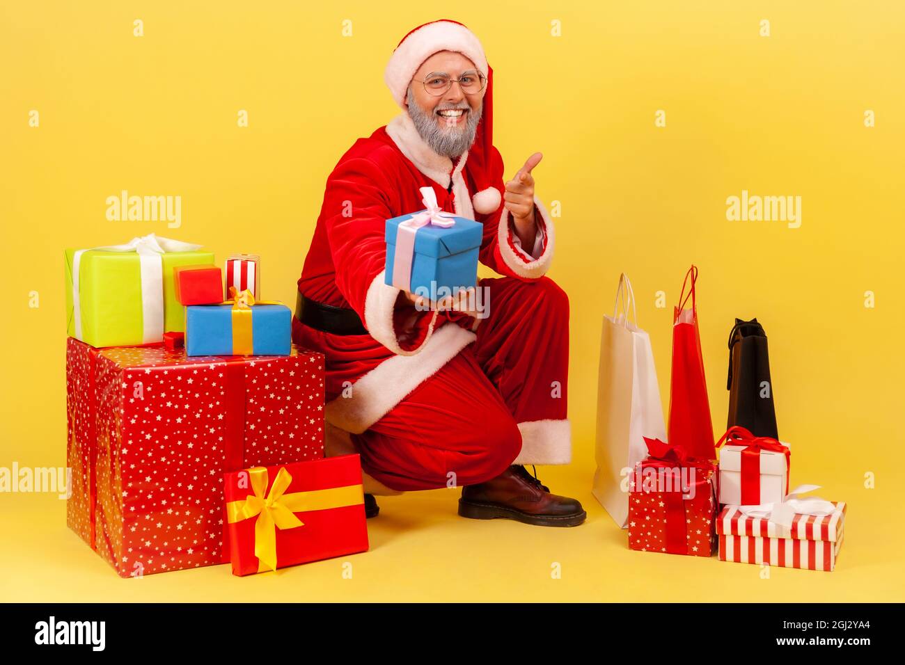Positive elderly man with gray beard wearing santa claus costume sitting near wrapped gift boxes, pointing to camera and giving present and smiling. I Stock Photo