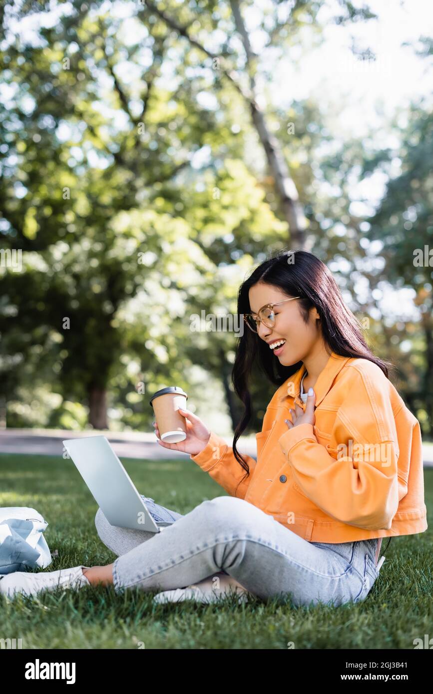 cheerful asian student with coffee to go touching chest while sitting on lawn with laptop Stock Photo