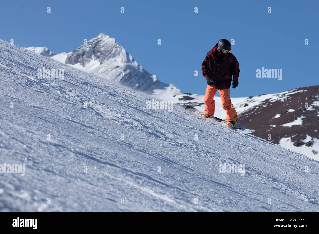 a man riding a snowboard down a snow covered slope on a sunny day with snowy mountains in the background wearing a helmet Stock Photo