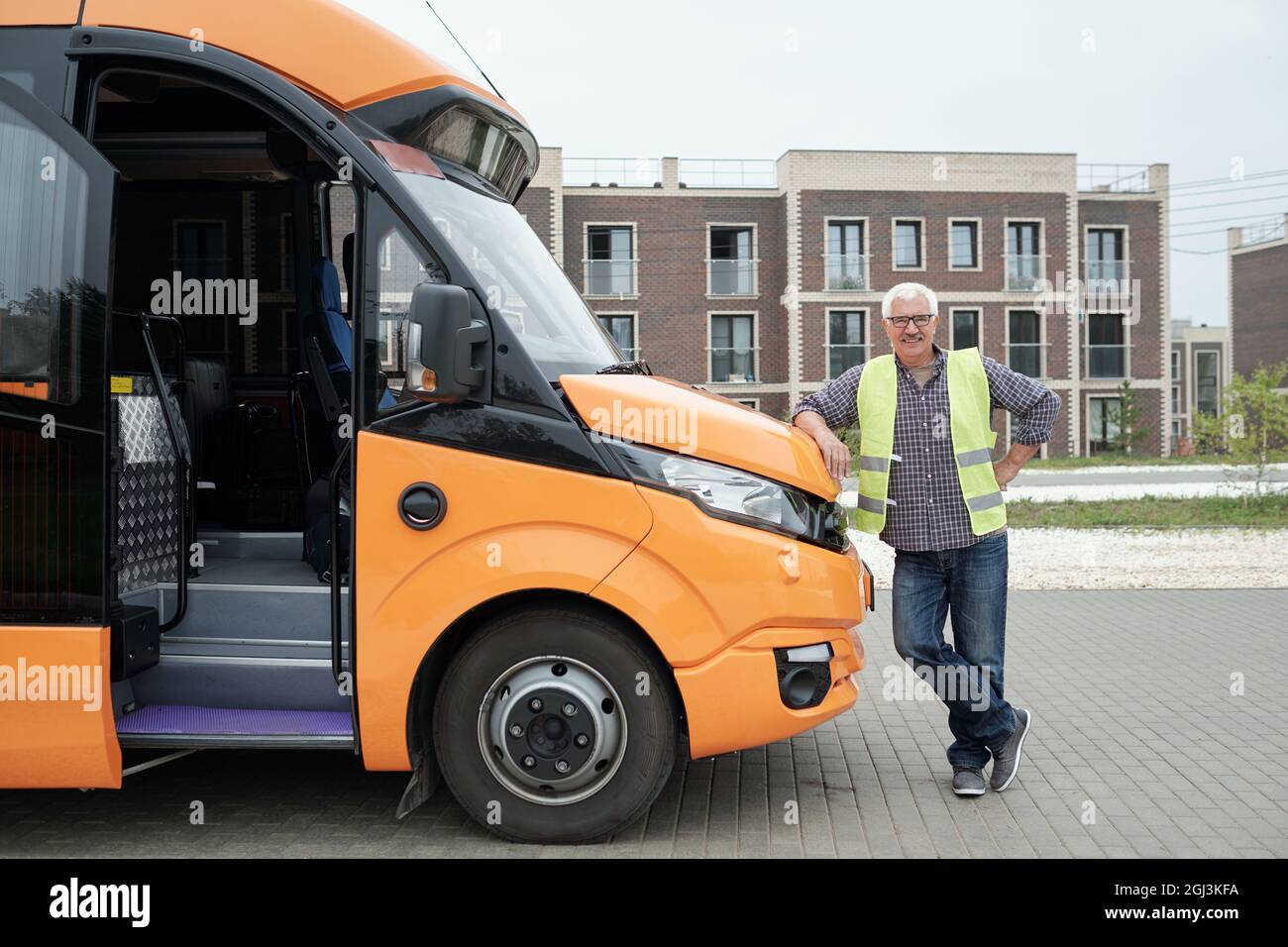 Smiling aged Caucasian bus driver in eyeglasses and green vest leaning on bonnet while waiting for passengers Stock Photo