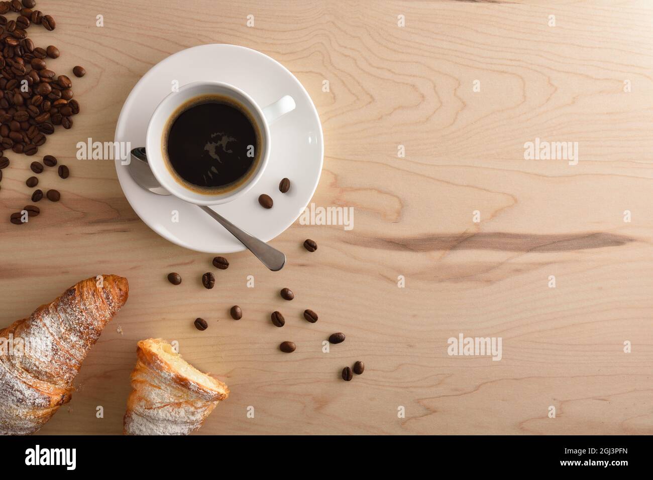 Porcelain white cup with hot coffee, croissant and beans on brown wooden table at breakfast. Top view. Horizontal composition. Stock Photo