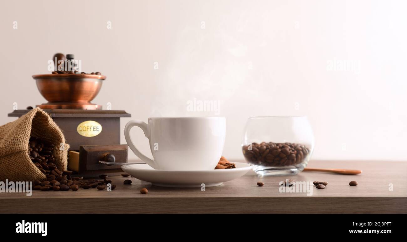 Cup of hot coffee with sack full of beans and manual grinder on wooden table and white isolated background. Front view. Horizontal composition. Stock Photo