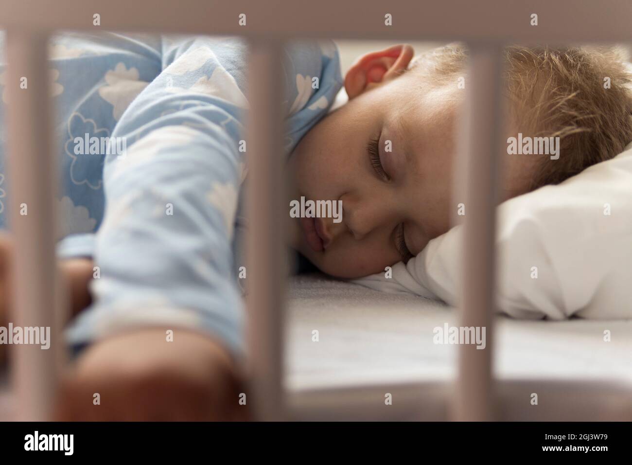 Cute cheerful little 2-3 years preschool baby boy kid sleeping sweetly in white crib during lunch rest time in blue pajama with pillow at home Stock Photo