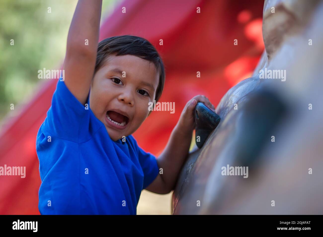 A cute boy shouting for joy as he climbs to the top of an outdoor rock climbing wall for kids. Stock Photo