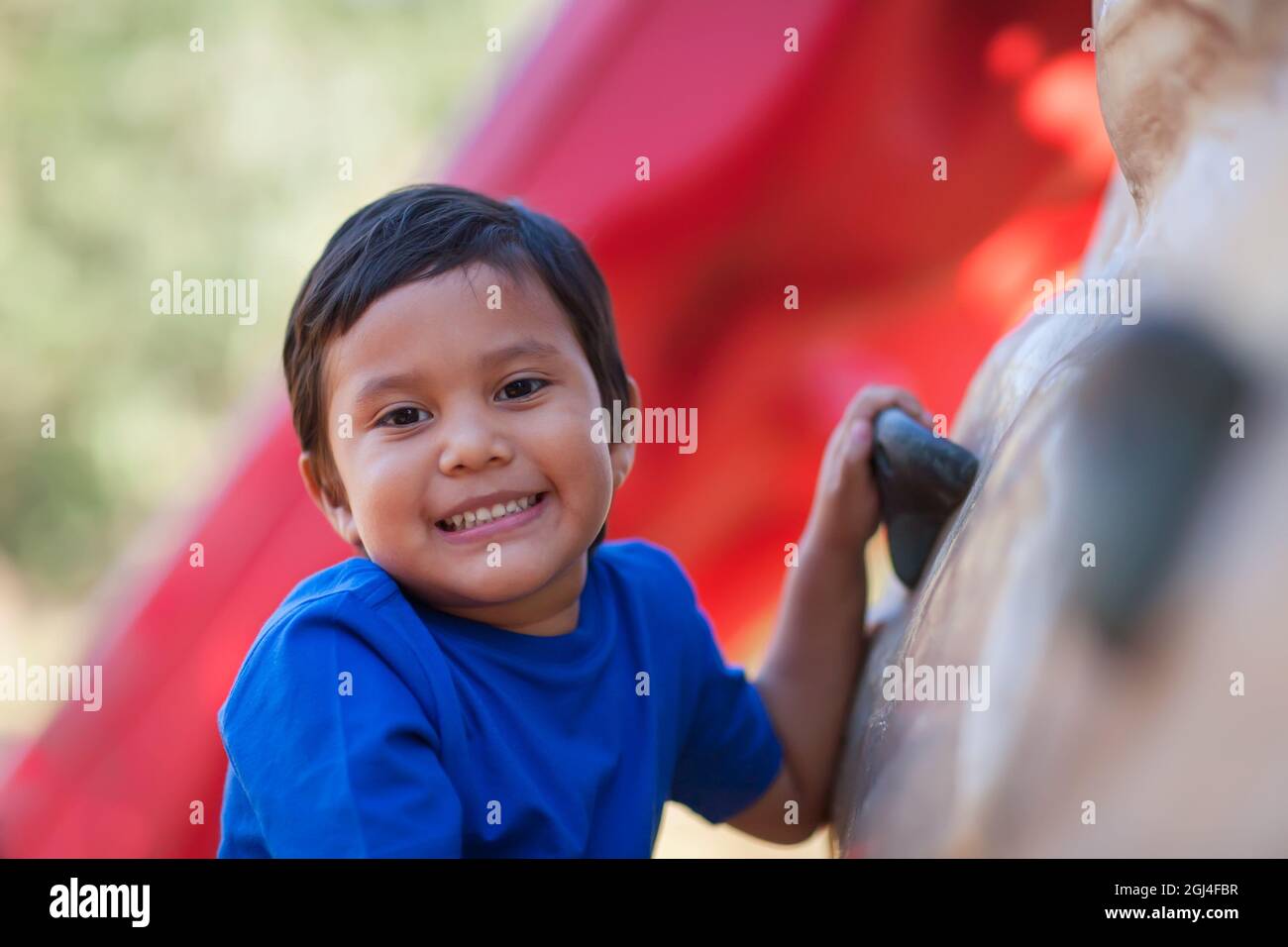 5 year old boy smiles while holding on to a kids outdoor rock climbing wall. Stock Photo