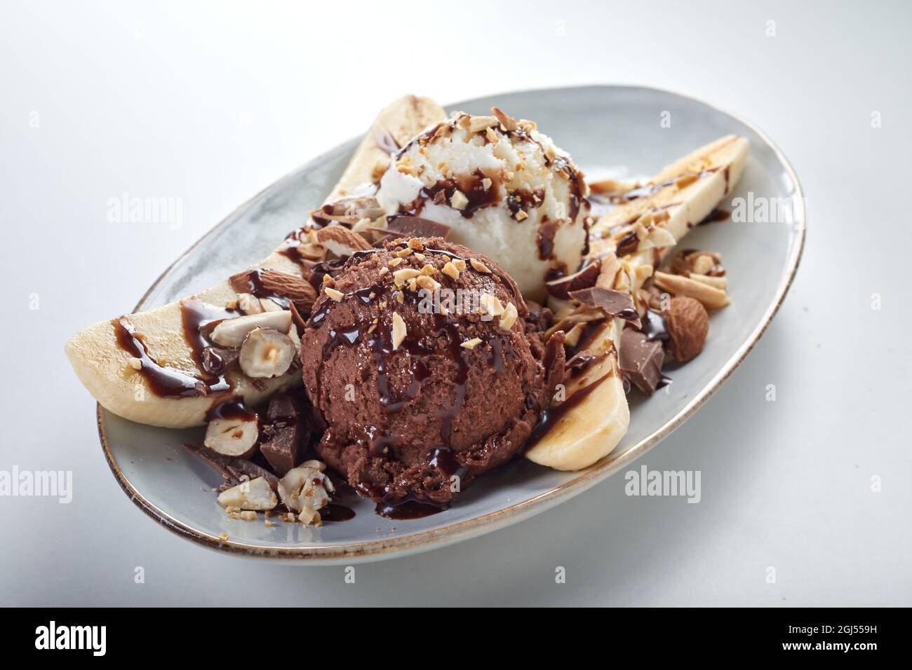 High angle of cold ice cream balls placed between banana slices garnished with chocolate syrup and hazelnuts on white background Stock Photo
