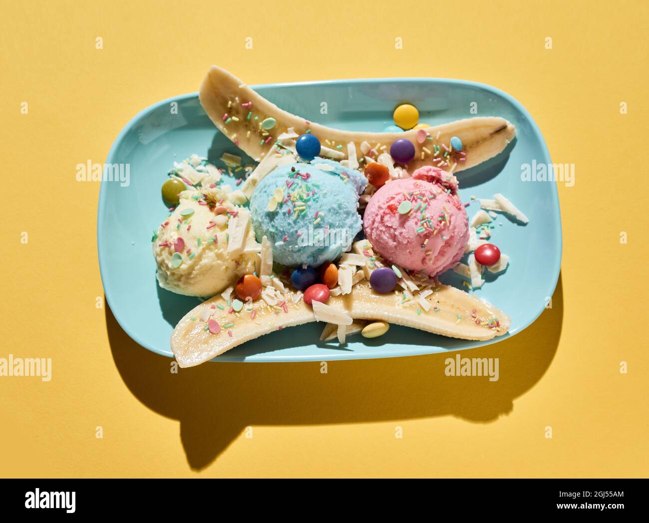 Top view of delicious banana split dessert with colorful ice cream scoops decorated with dragee and sprinkles served on blue plate on yellow backgroun Stock Photo