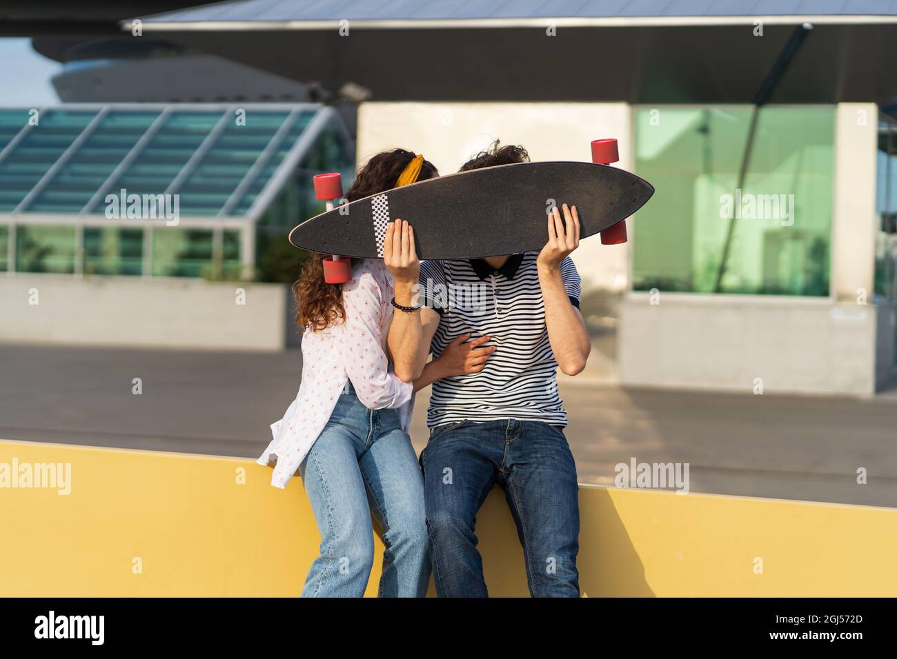 Joyful couple kiss hiding above longboard. Playful man and woman in love together hold skateboard Stock Photo