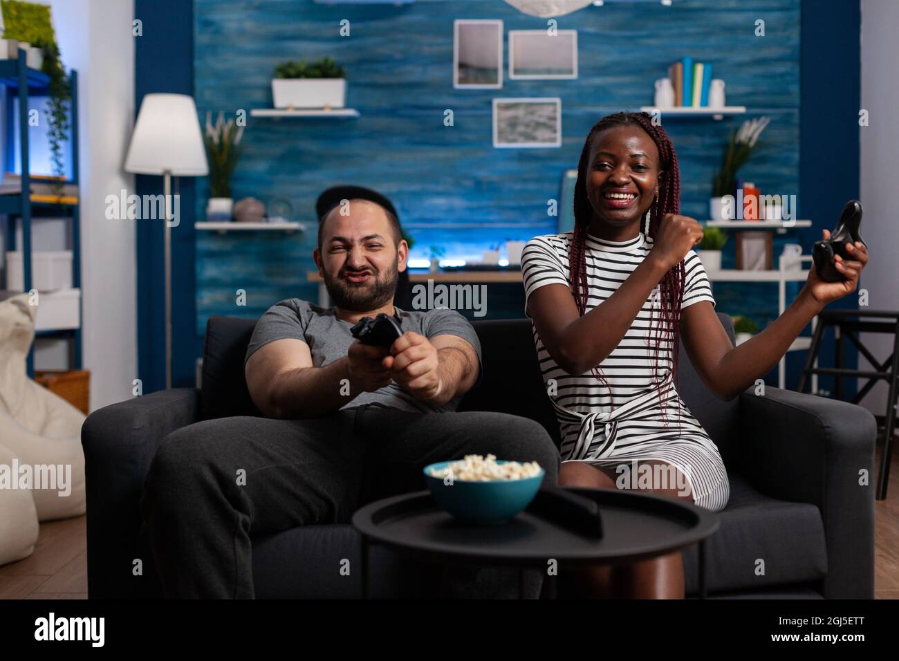 Cheerful interracial people winning video game on TV while playing with console and joysticks. Mixed race couple using technology for entertainment, having fun on sofa using controllers Stock Photo
