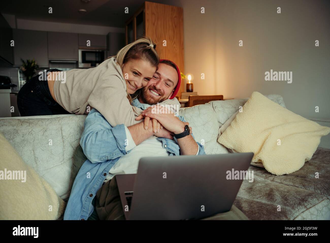 Caucasian happy couple sitting on couch showing love while hugging and cuddling while on video call using laptop Stock Photo
