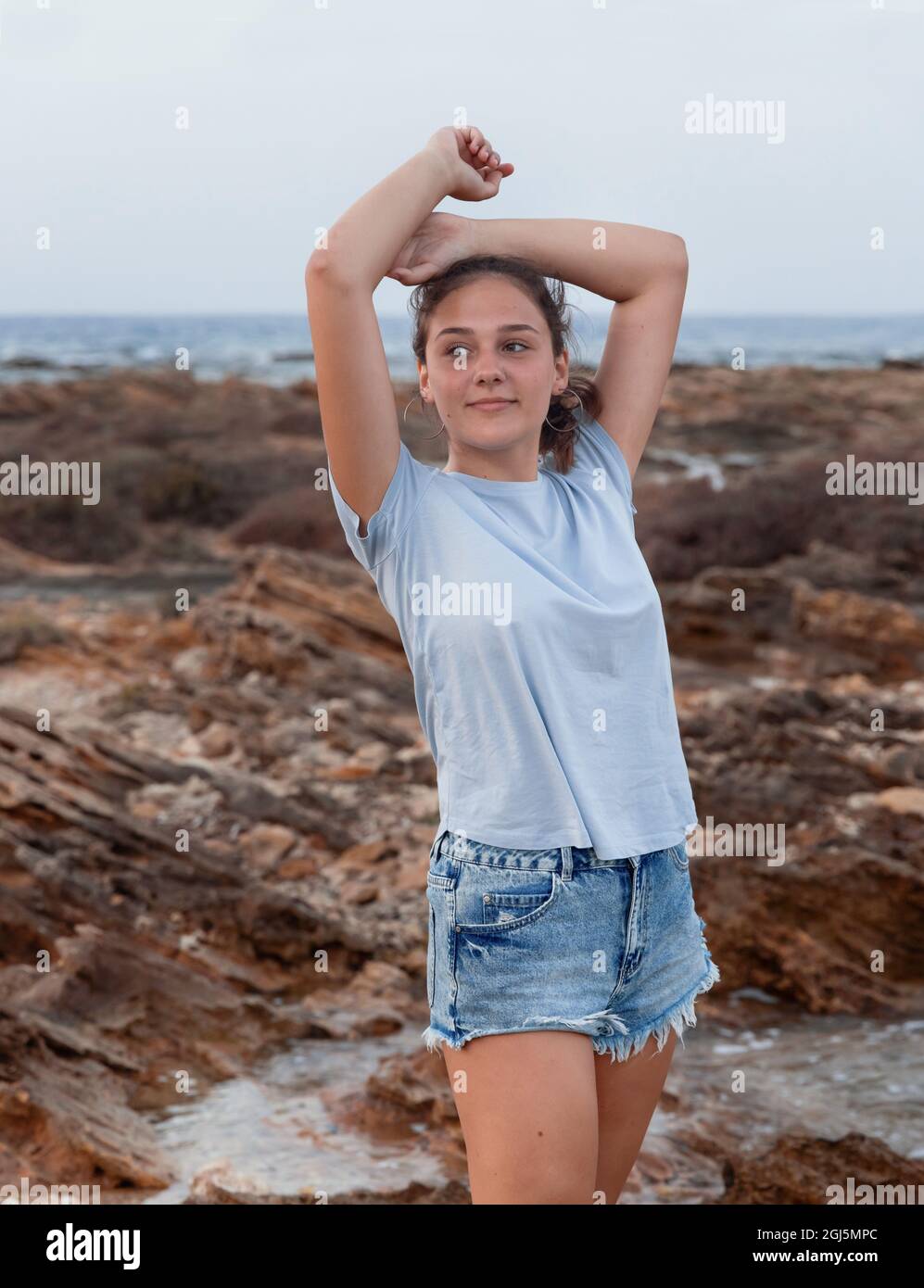Teenage girl standing with heads over head on cliff by the sea at sunset wearing light blue t-shirt, jeans shorts. Three quarter length shot. T-shirt Stock Photo