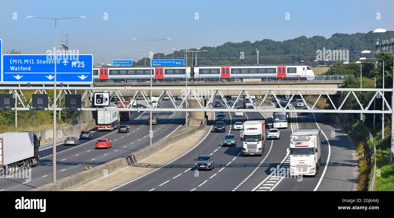 M25 motorway gantry signs railway bridge & Greater Anglia passenger train crossing above road traffic junction 28 for A12 Brentwood Essex England UK Stock Photo