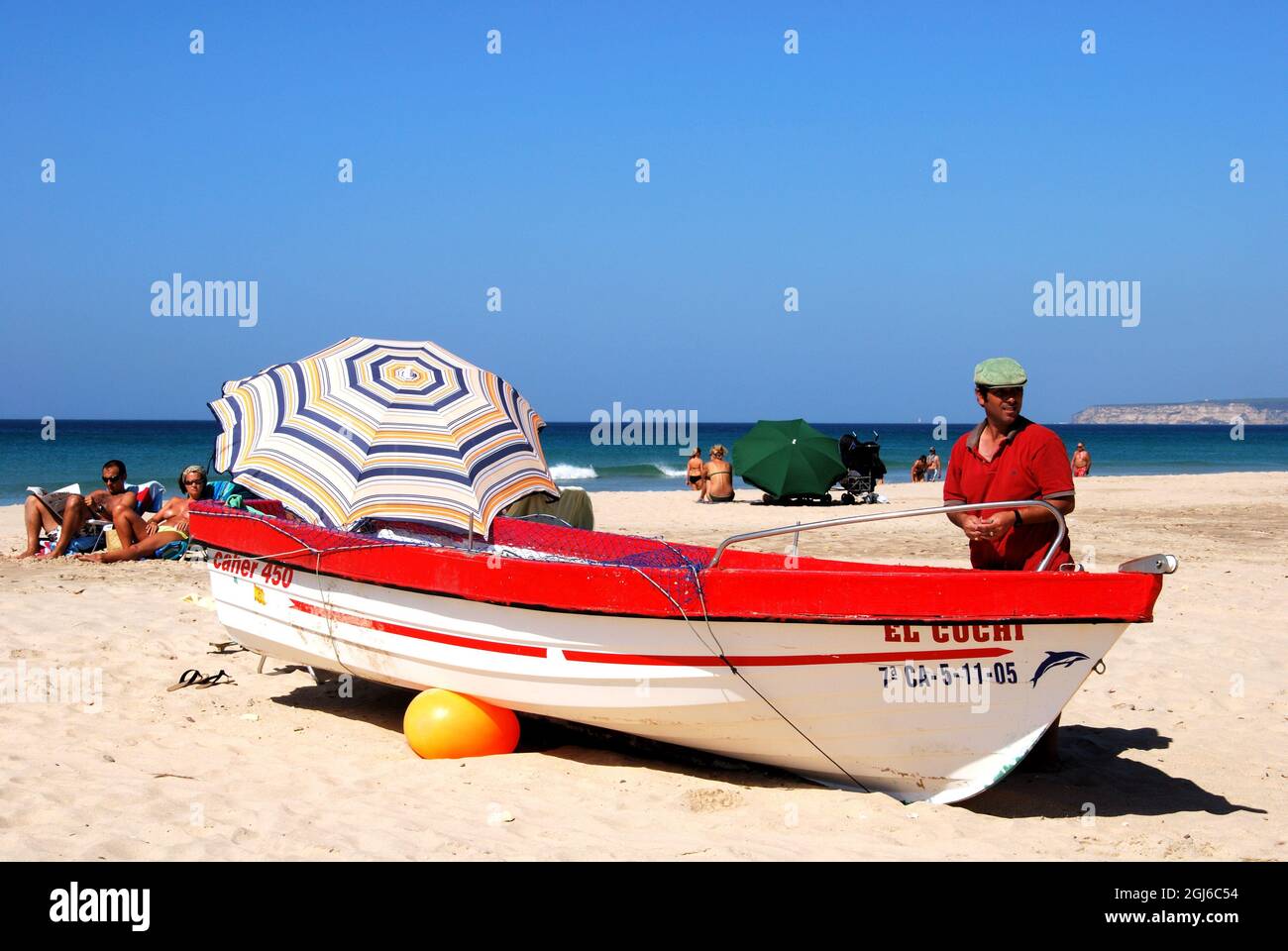 Spanish man standing by his traditional red and white wooden fishing boat on the beach with tourist enjoying the sunshine, Zahara de los Atunes, Spain Stock Photo