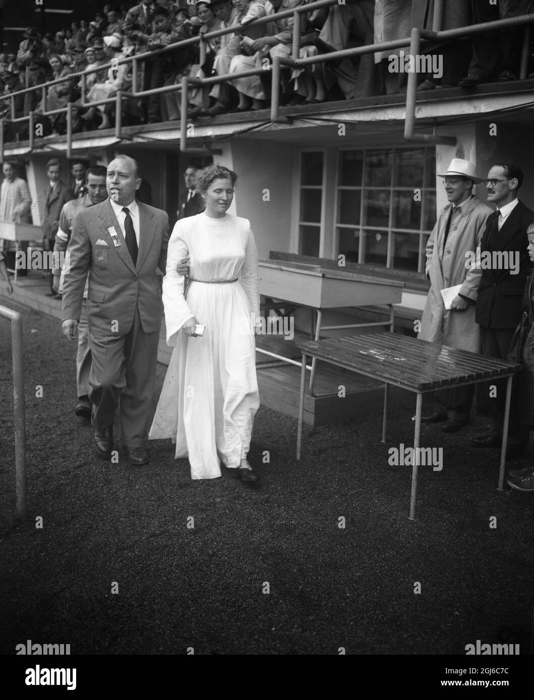 An Olympic official escorts student , Barbara Pleyer ( dressed in white ) , from the Olympic stadium in Helsinki during the official opening ceremony . She had tried to gate - crash the oath taking at the opening ceremony . 19th July 1952 Stock Photo
