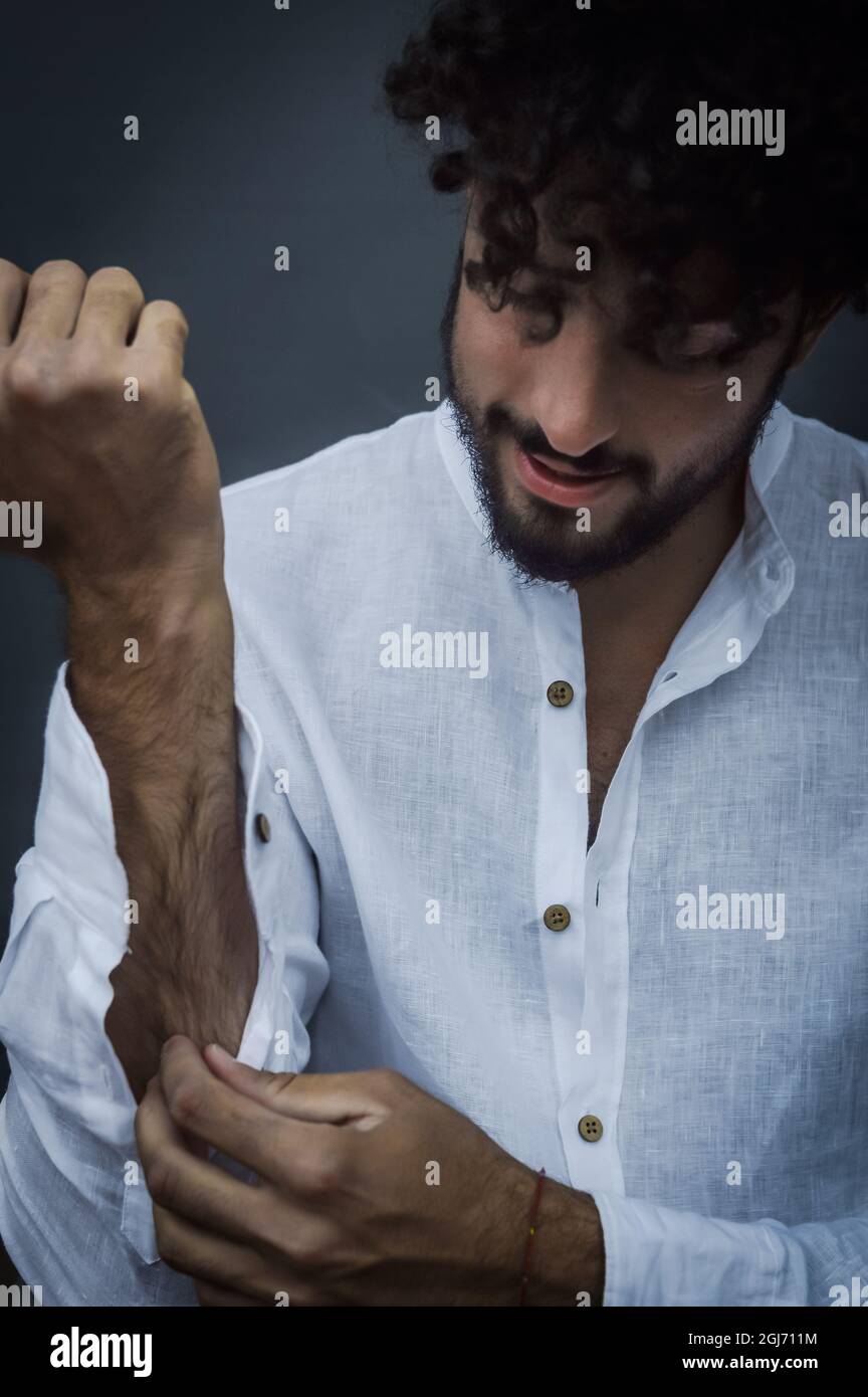 portrait of a young man with a beard and curly hair adjusting the sleeves of his white shirt while dressing Stock Photo